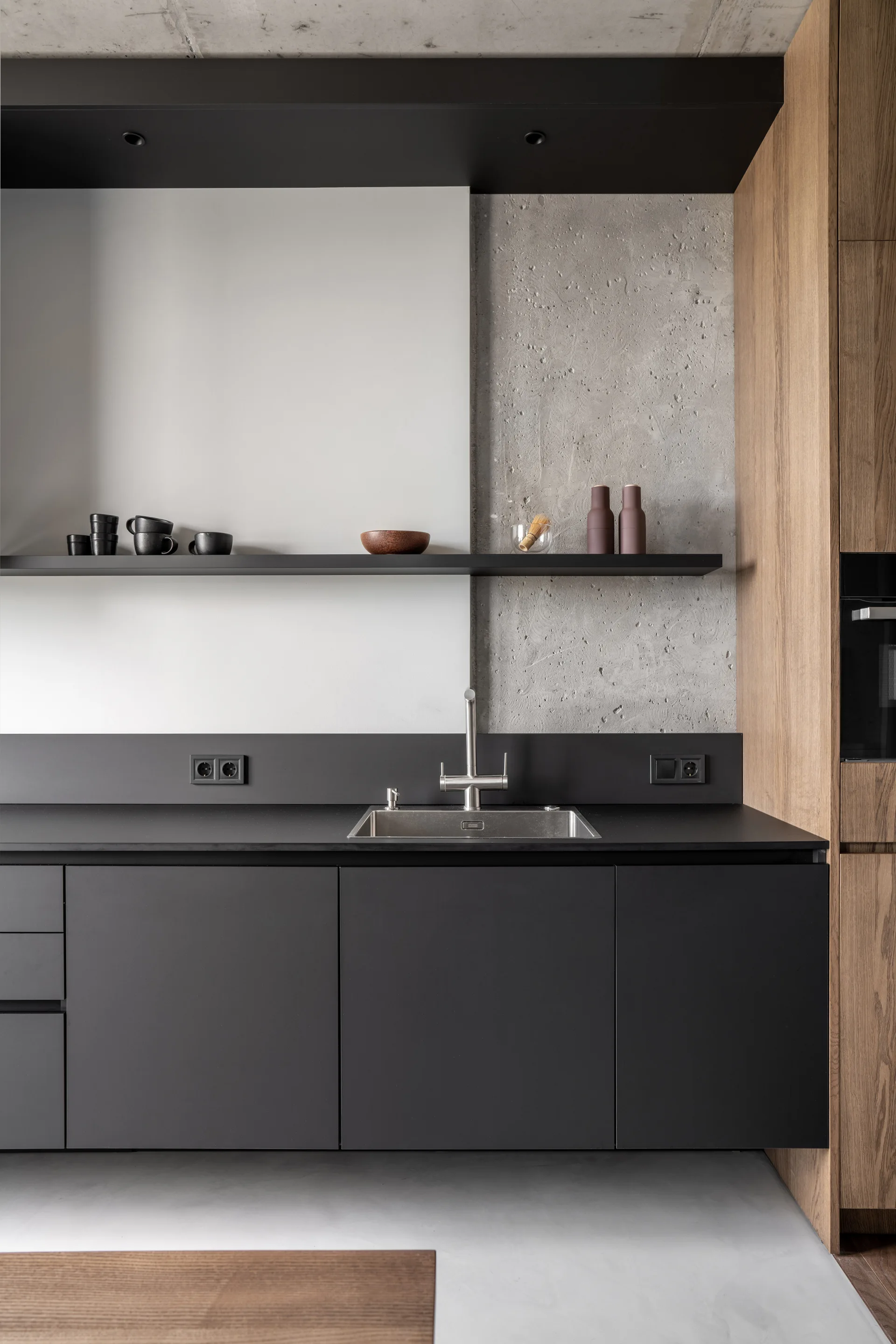 Minimalist kitchen features matte black cabinets, a stainless steel sink, a black floating shelf, and a textured concrete and smooth white backsplash.