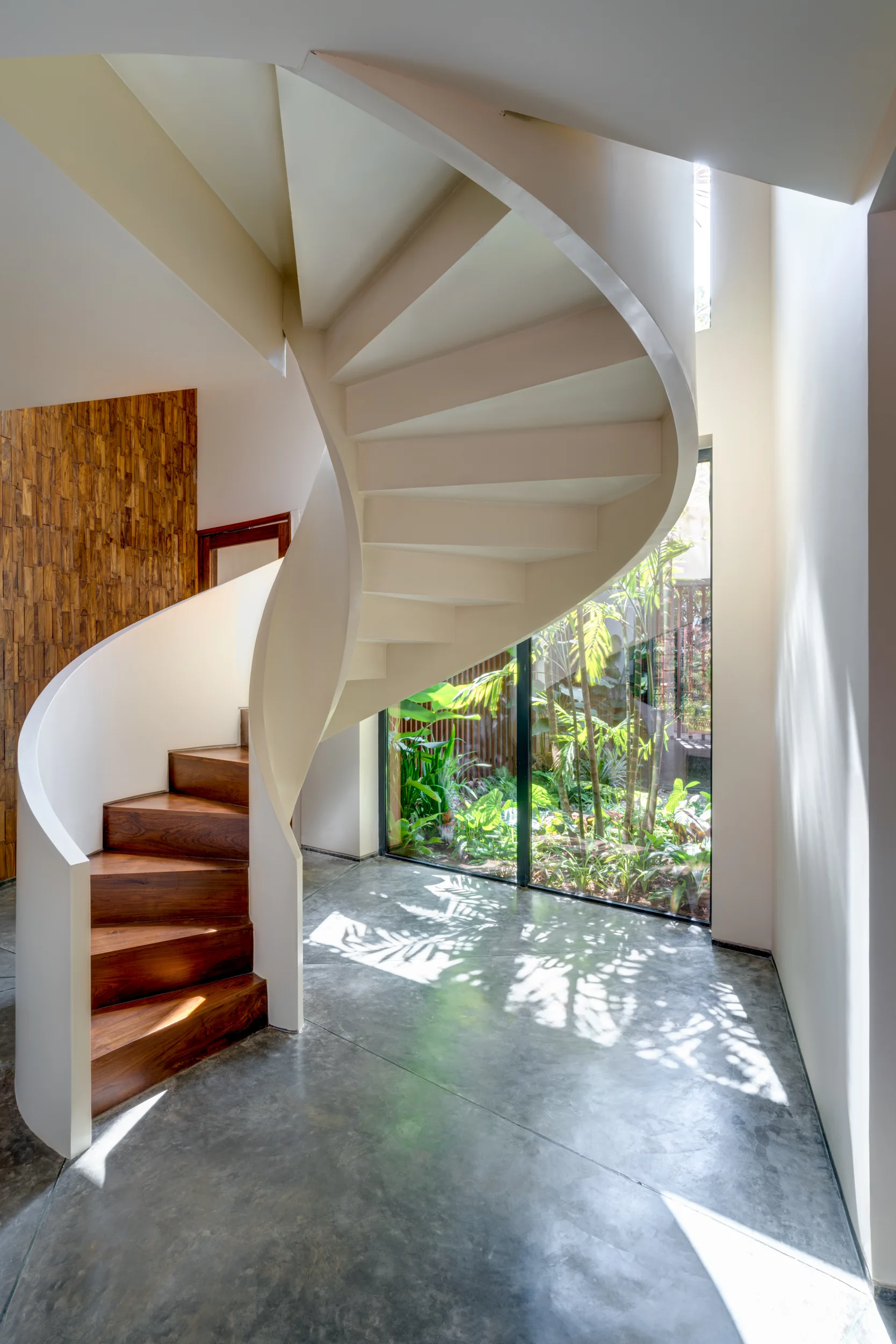 A modern white spiral staircase with polished wood treads stands on a grey concrete floor next to a large window overlooking a green garden.