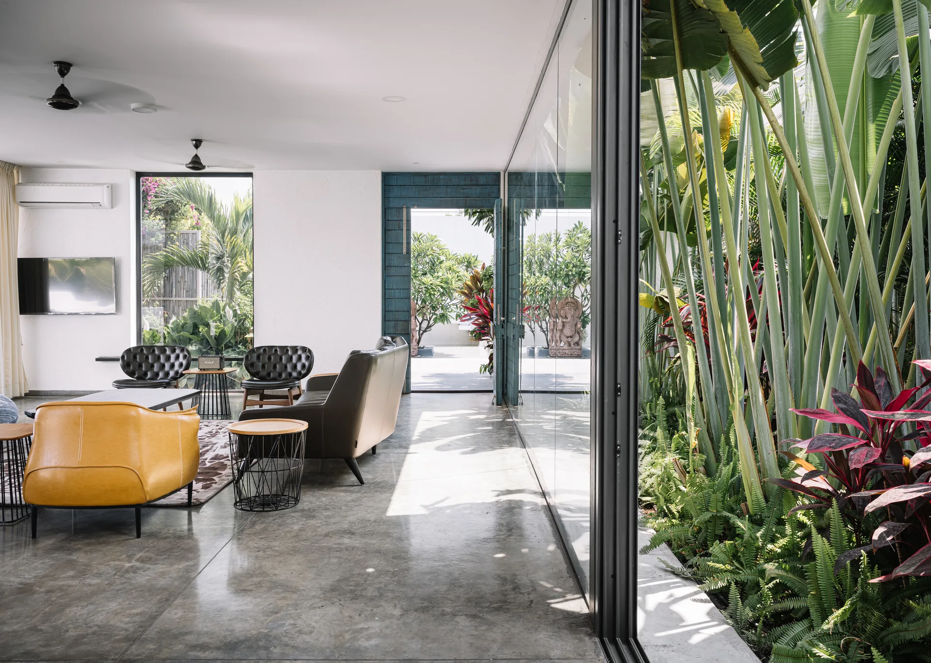 A modern living room with leather furniture and polished concrete floors features a large glass wall looking out at a tropical garden.