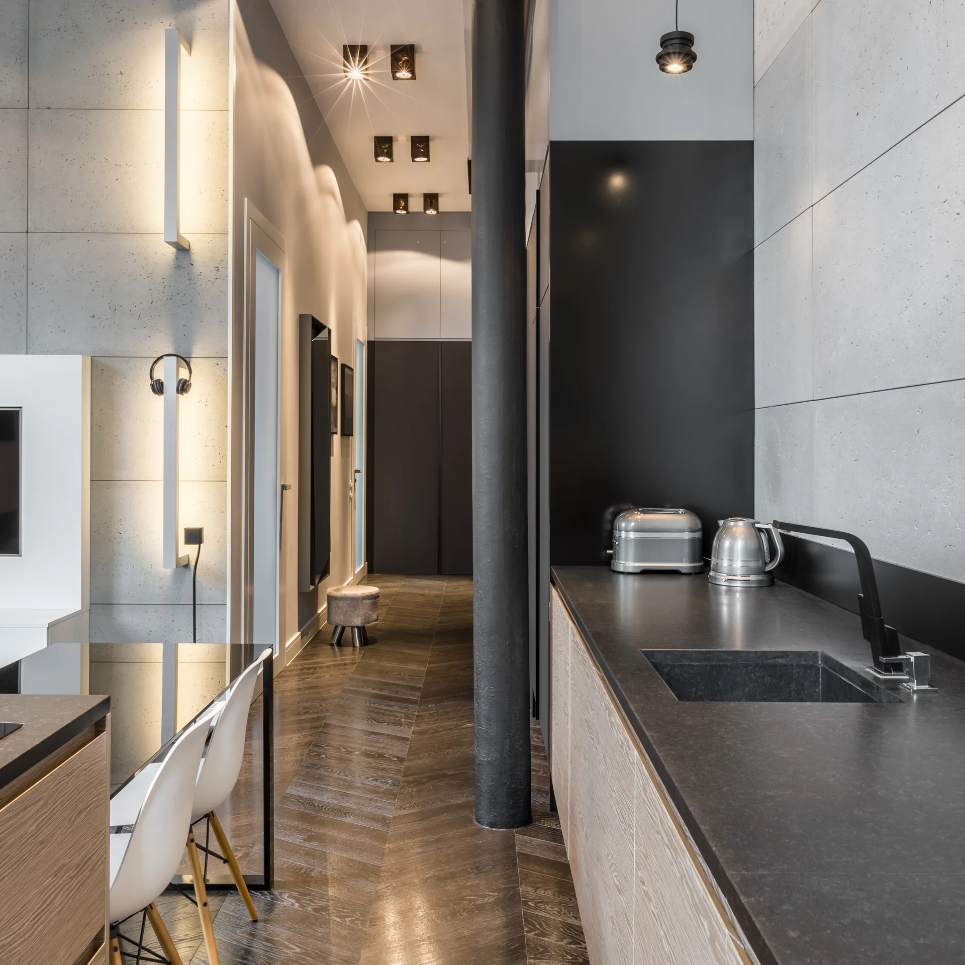 A modern open-plan kitchen featuring a dark countertop, light wood cabinetry, concrete-panel walls, and a central black structural column on herringbone floors.