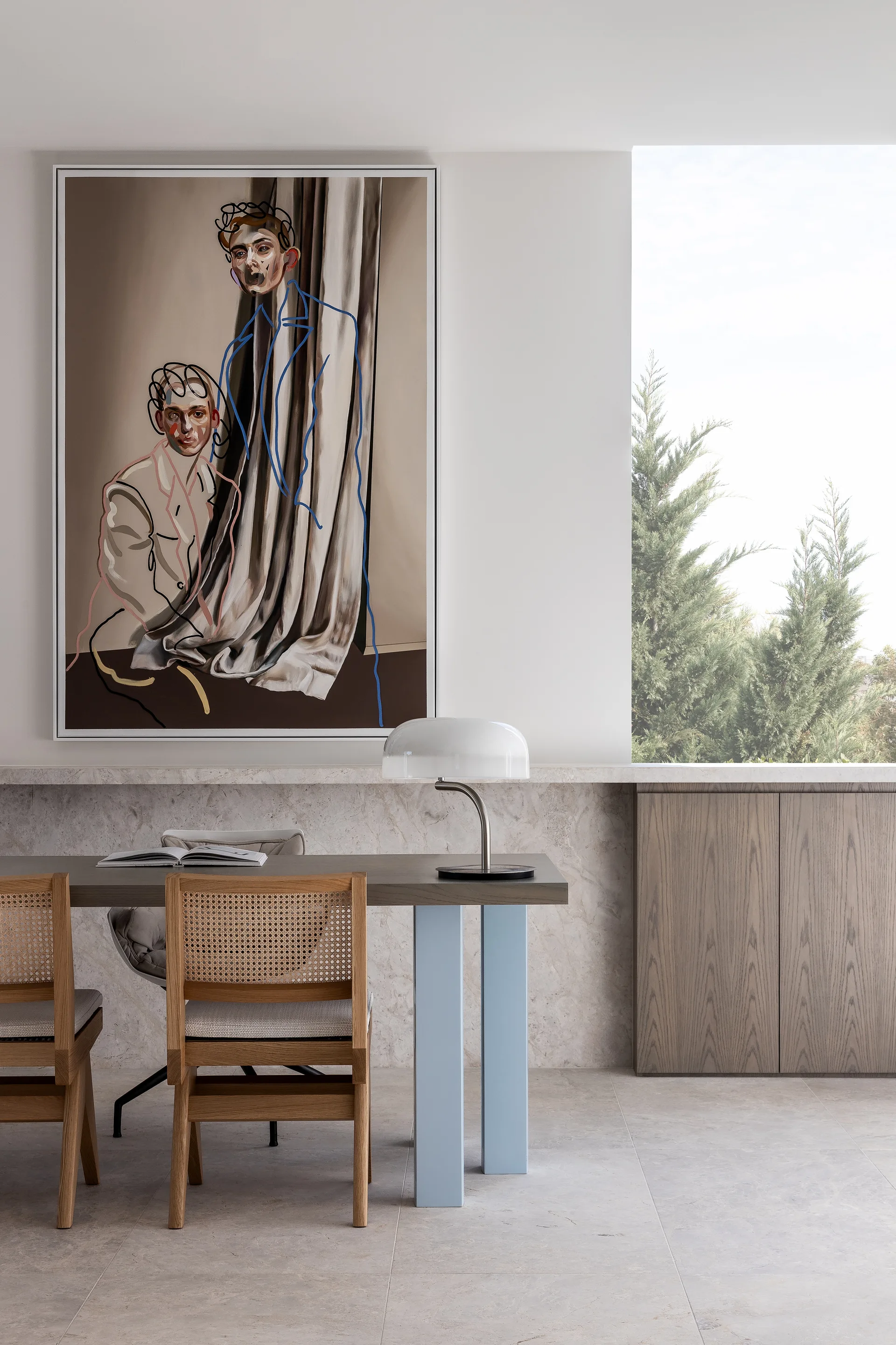 A contemporary dining area featuring a wooden table with blue legs, cane-back chairs, a large abstract painting, and a marble-clad lower wall.