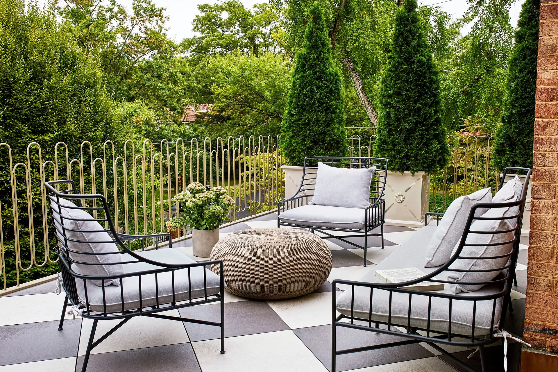 An outdoor patio features black metal armchairs and a sofa on a black and white checkerboard tiled floor, surrounded by a metal railing and tall green trees.