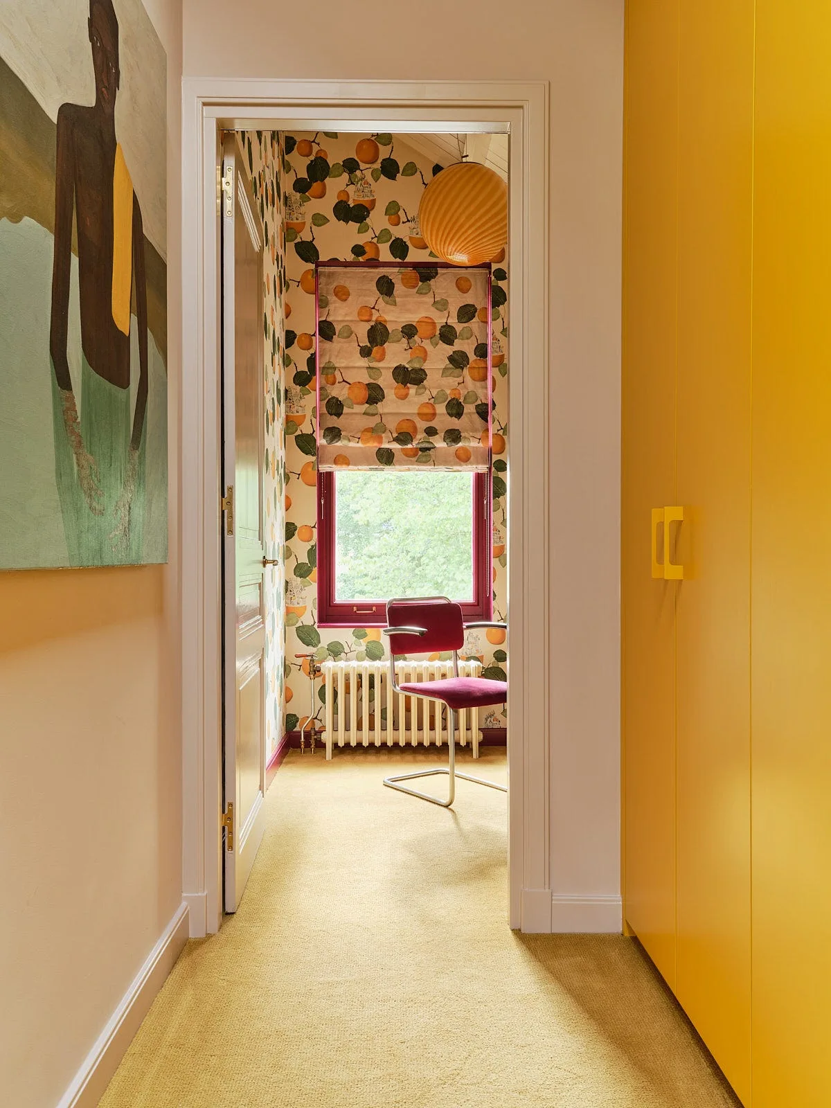 A yellow hallway with tall yellow cabinets leads to a small room featuring citrus-patterned wallpaper and a burgundy cantilever chair.