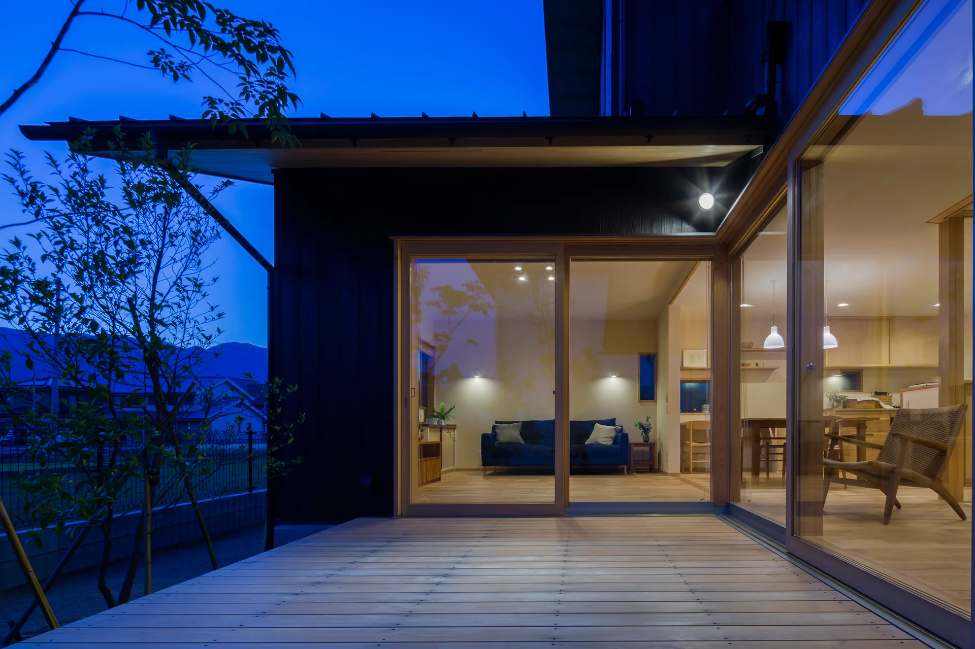 A modern dark wood house exterior features a light wood deck and large sliding glass doors revealing a warmly lit interior living room.