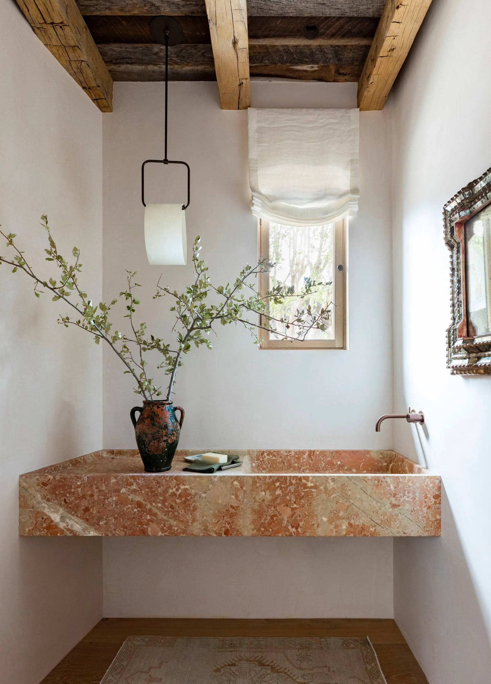 A powder room features a monolithic peach stone sink, a copper wall-mounted faucet, rough timber ceiling beams, and a small window with a linen shade.