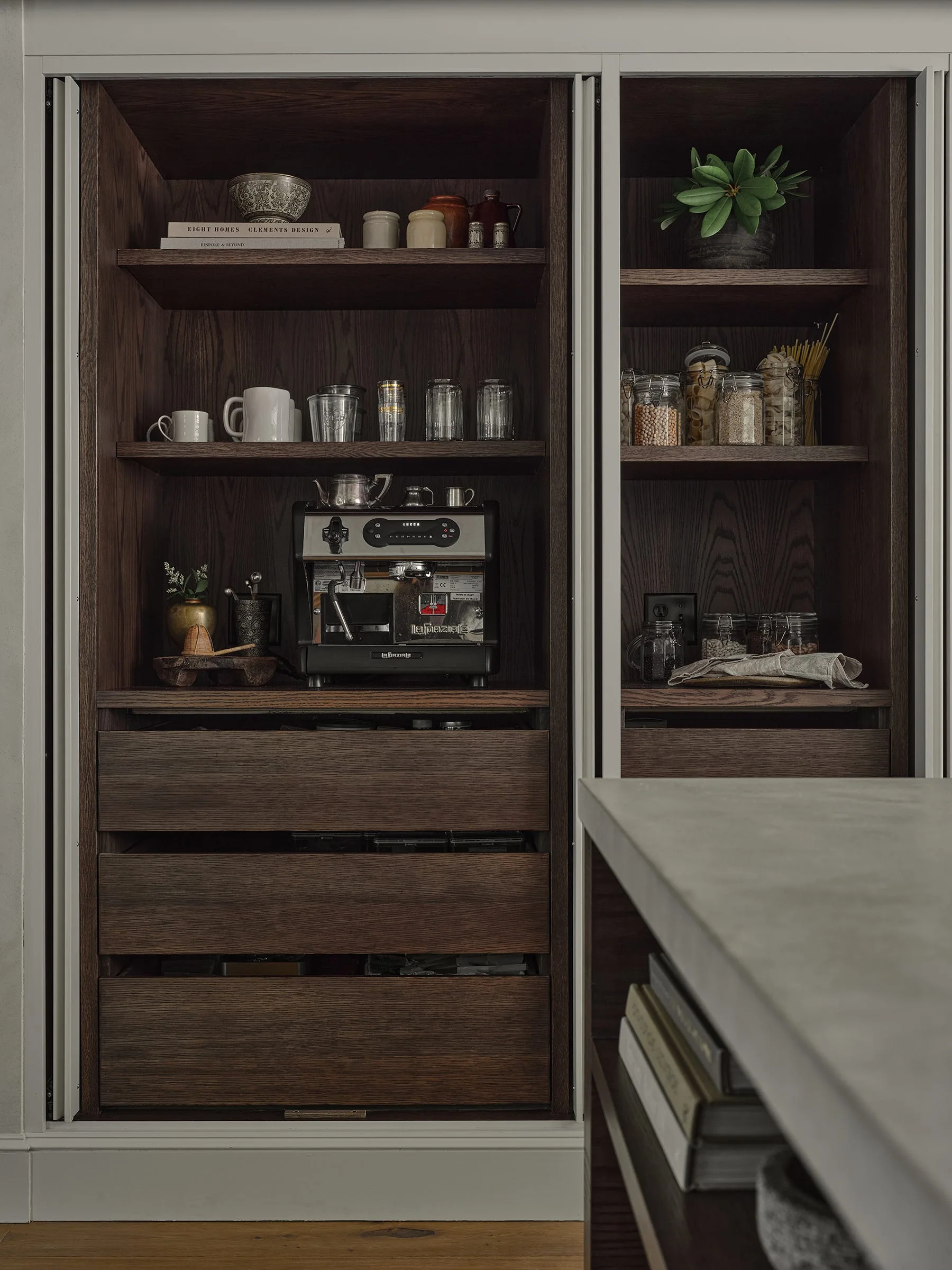 A dark wood built-in pantry cabinet features open shelves with an espresso machine, glass storage jars, and wooden drawers below.