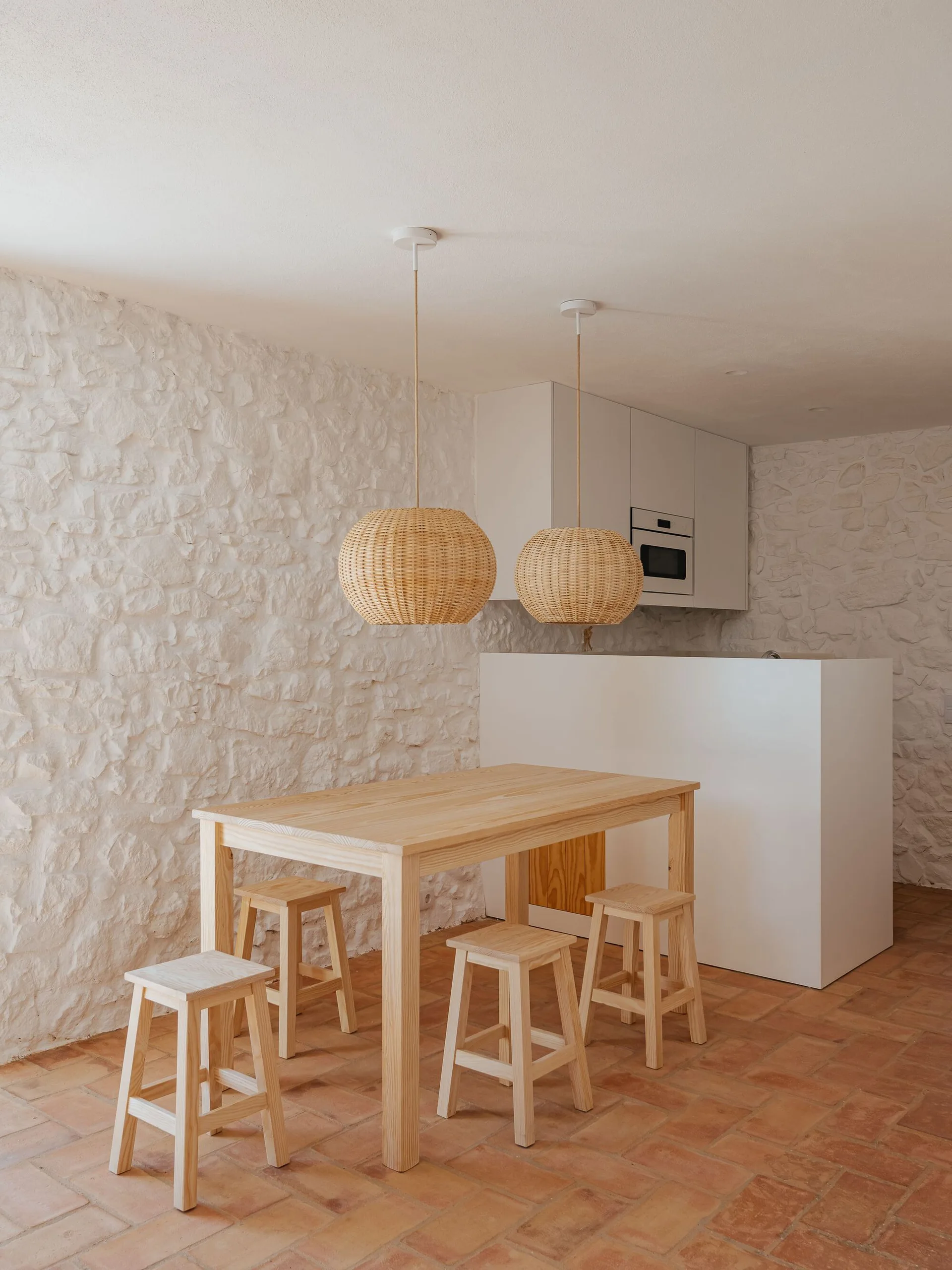 A light wood dining table and stools stand on a terracotta tiled floor beneath two woven rattan pendant lights and textured white stone walls.