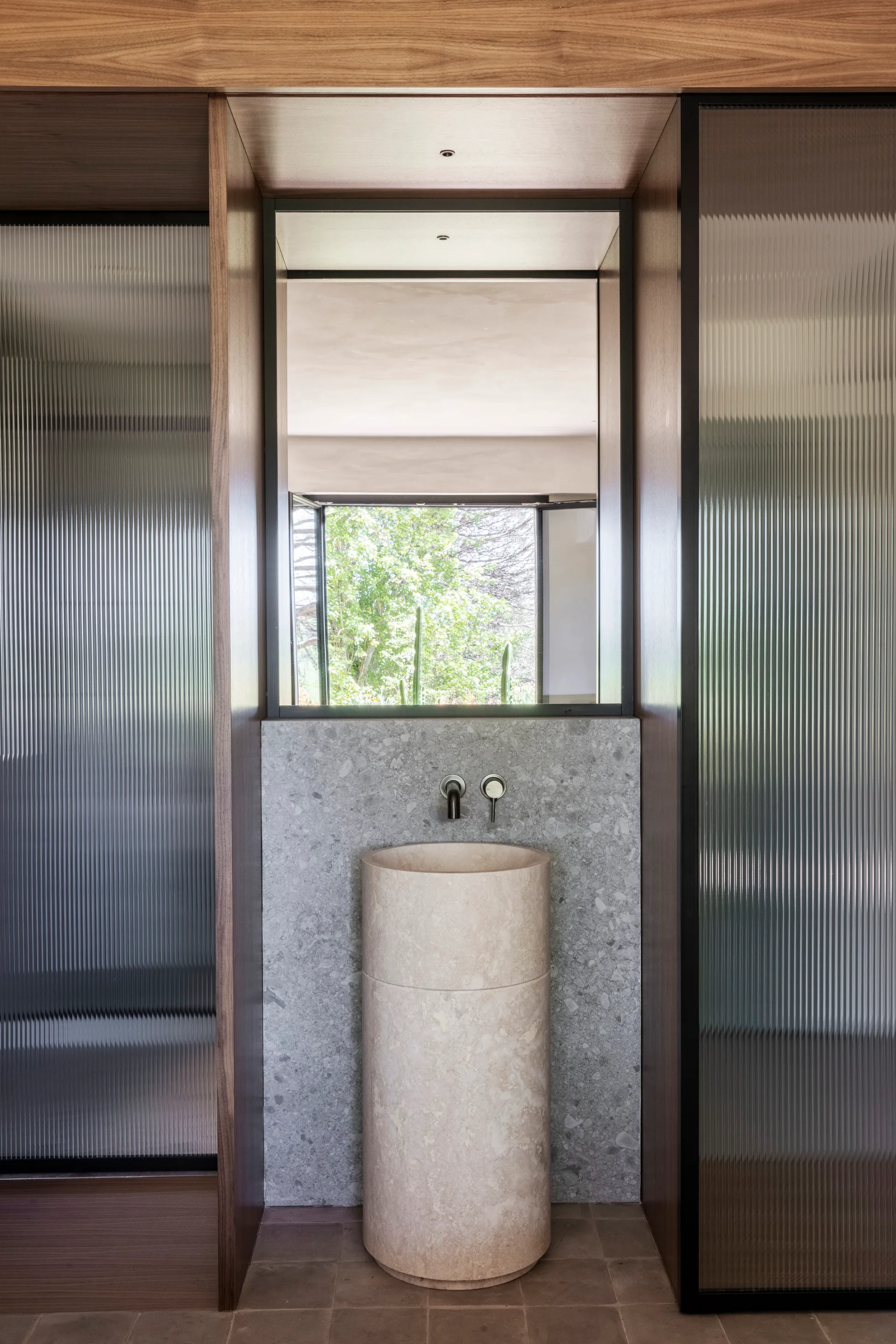 A modern powder room featuring a cylindrical beige stone pedestal sink set against a grey stone backsplash flanked by fluted glass partitions.