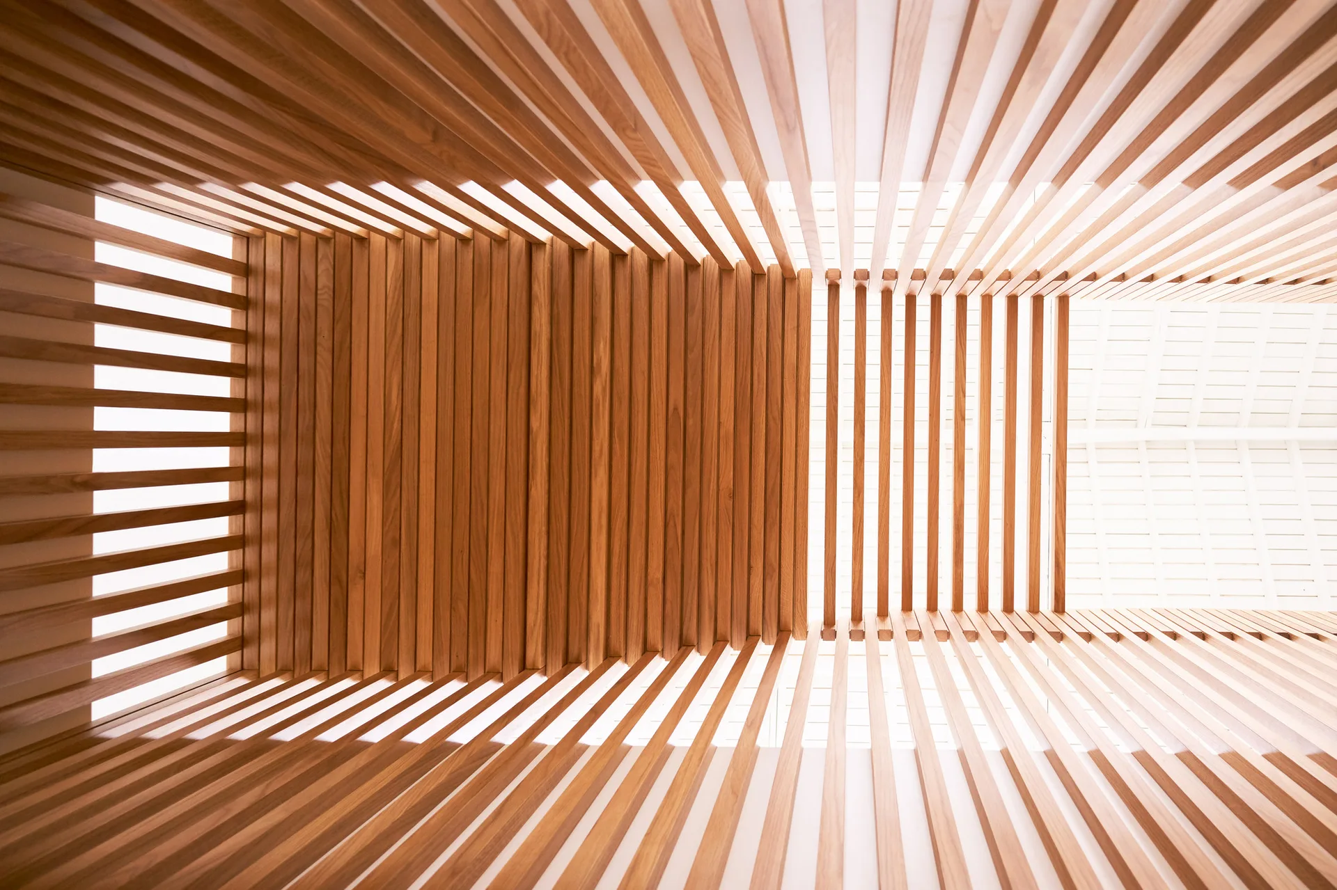 A low-angle view of a modern ceiling featuring parallel wooden slats with soft, diffused light filtering through the open gaps.