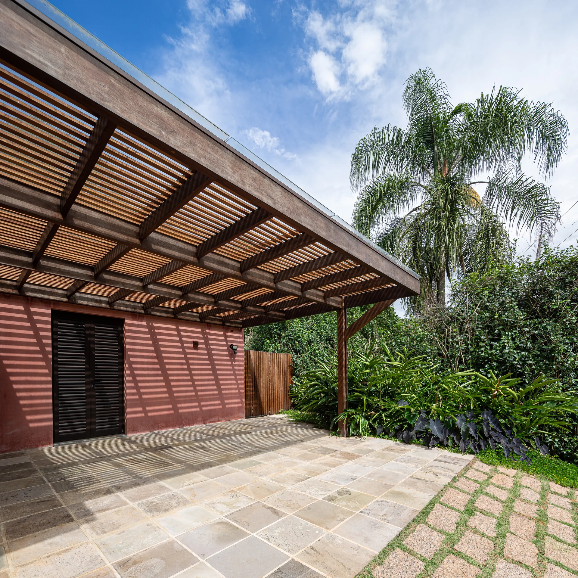 A wooden slatted pergola casts striped shadows on a stone tile patio next to a terracotta stucco wall with a dark slatted door and lush green garden.