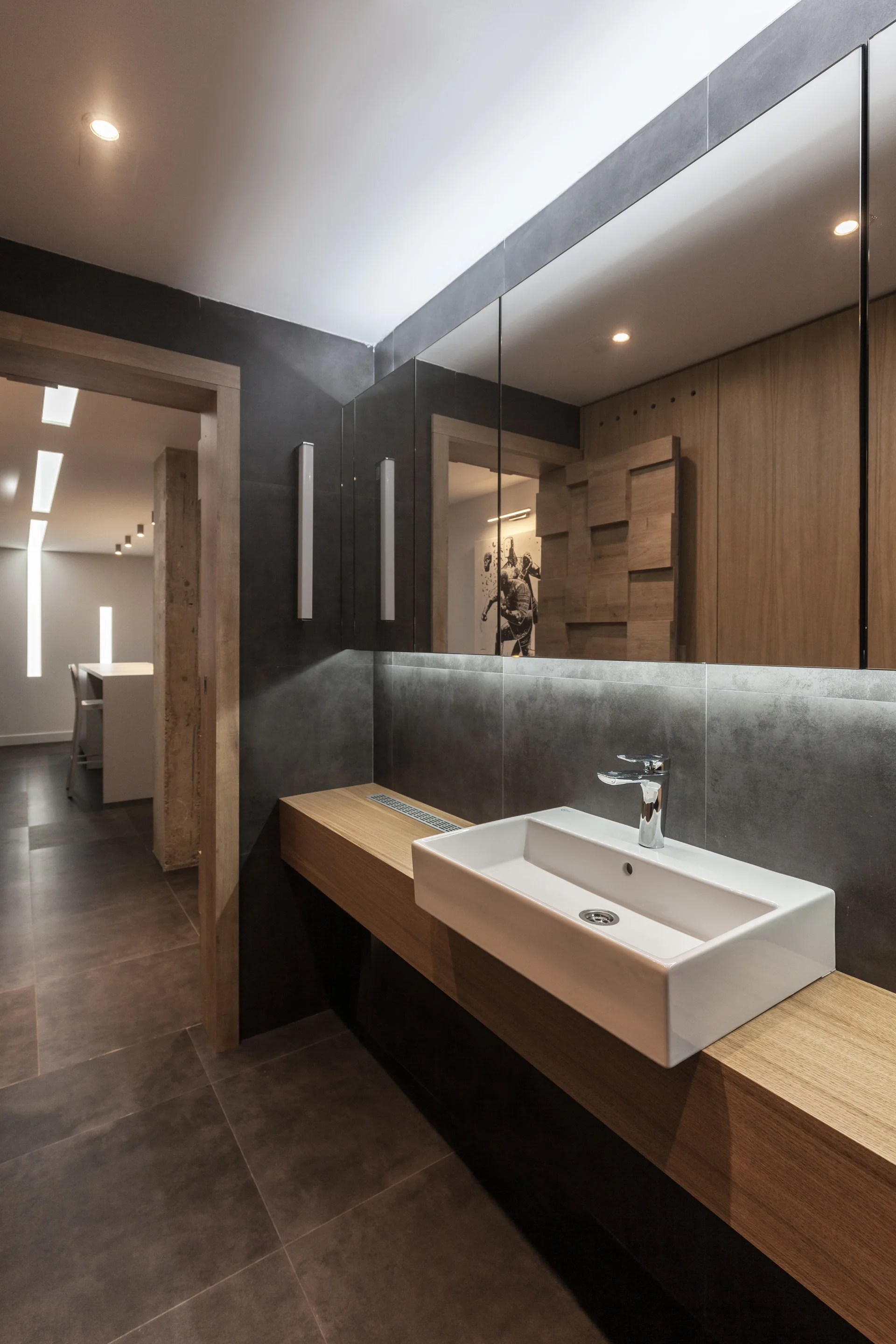 A modern bathroom vanity features a white ceramic vessel sink on a floating wood counter against dark grey tiled walls beneath a large mirror.