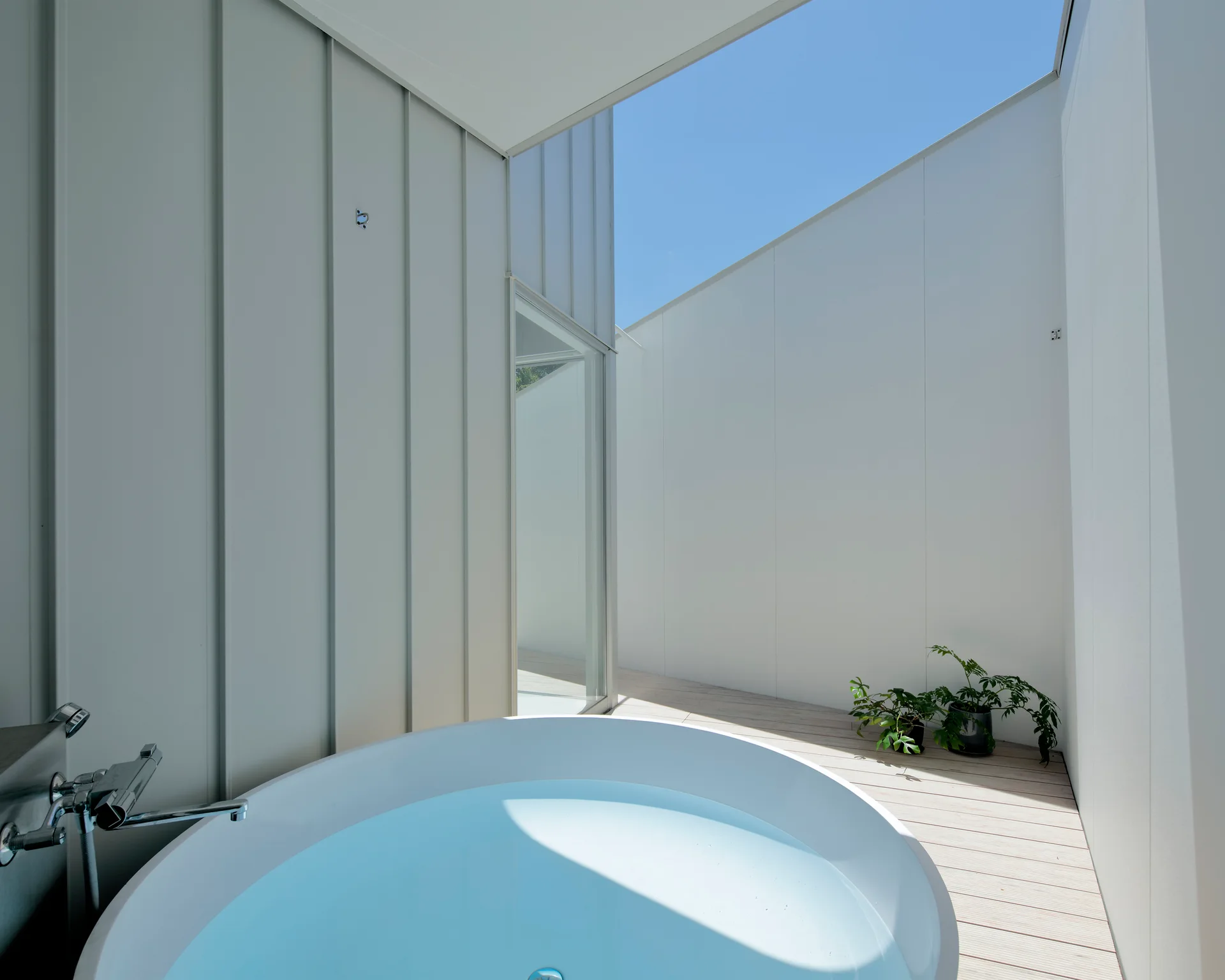 A circular white soaking tub sits on a light wood deck surrounded by tall off-white paneled walls under an open blue sky.