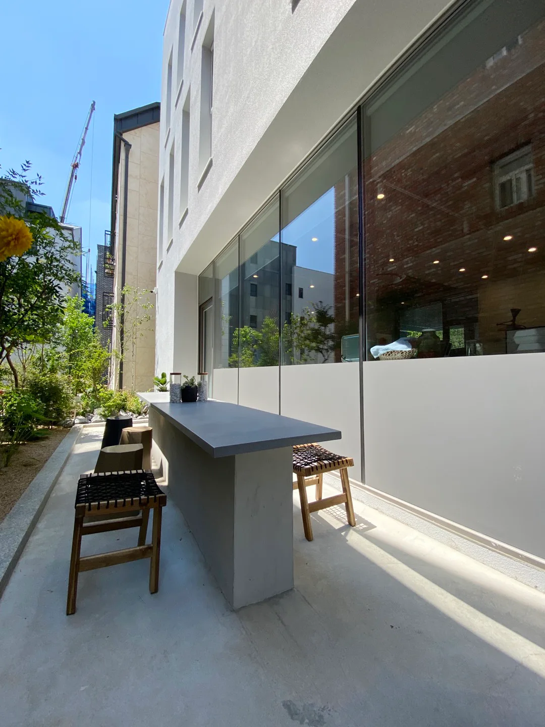 A narrow concrete patio features a long grey bar counter with woven wood stools alongside a large glass window and a small garden.