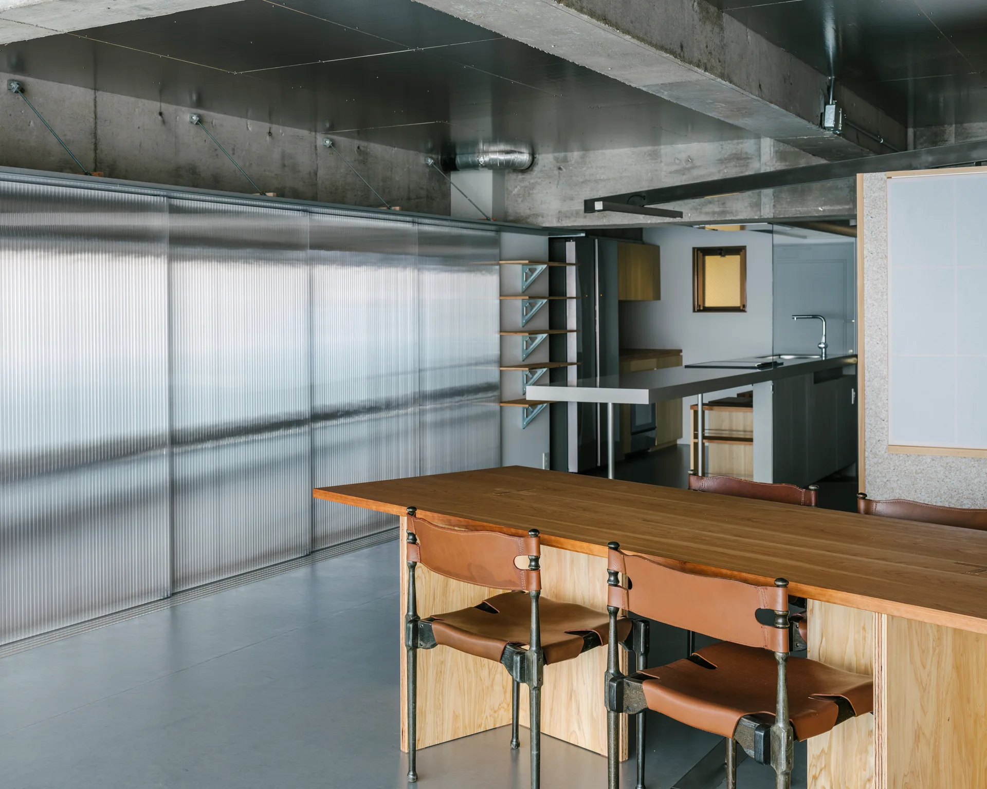 A wooden dining table with leather chairs sits in an industrial interior featuring a corrugated plastic sliding partition and exposed concrete walls.