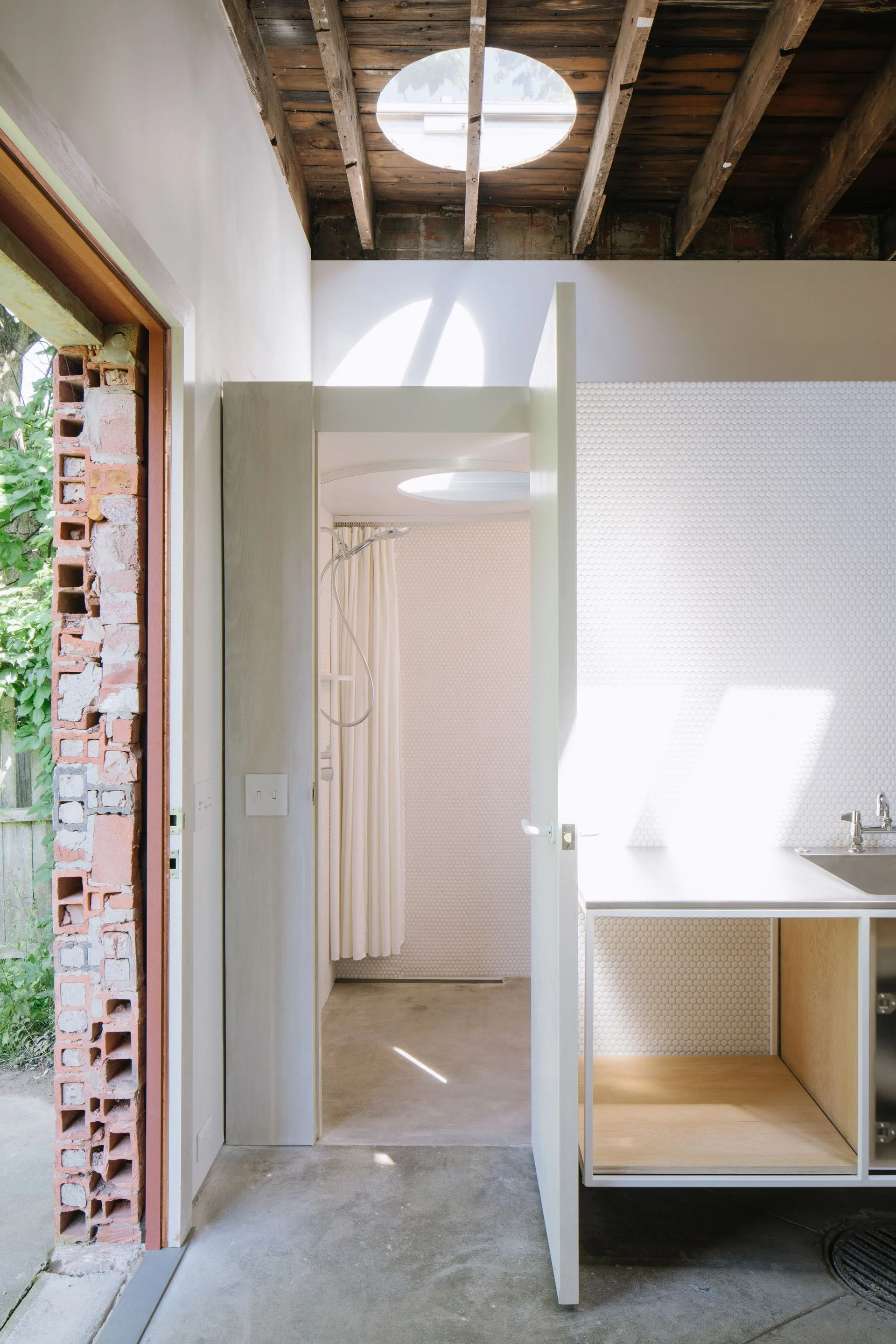 A minimalist bathroom featuring a circular skylight in an exposed wood ceiling, white penny tile walls, concrete flooring, and an open wood vanity.