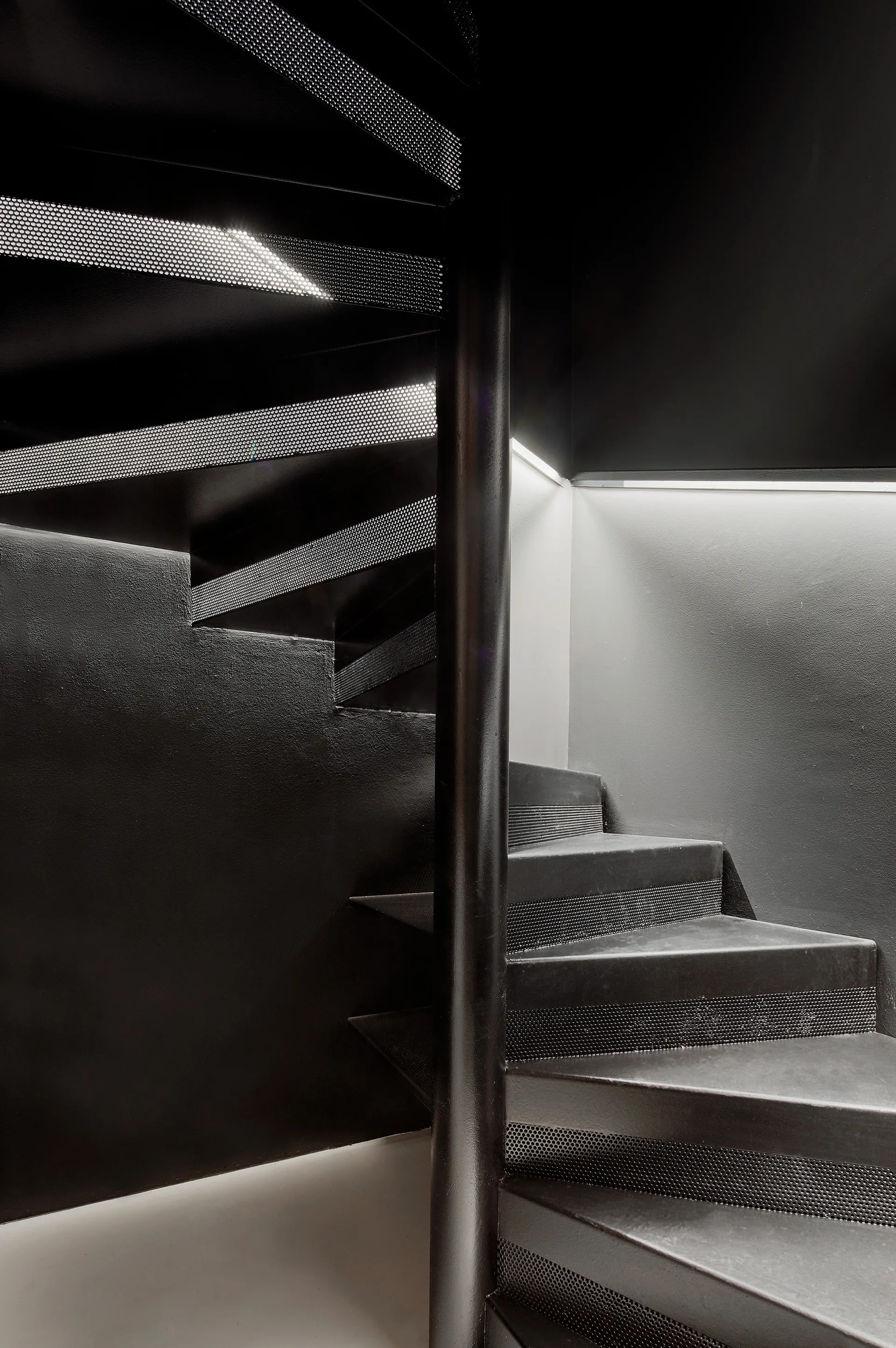 A modern matte black steel spiral staircase with perforated metal treads lit by integrated under-step strip lighting against a dark wall.