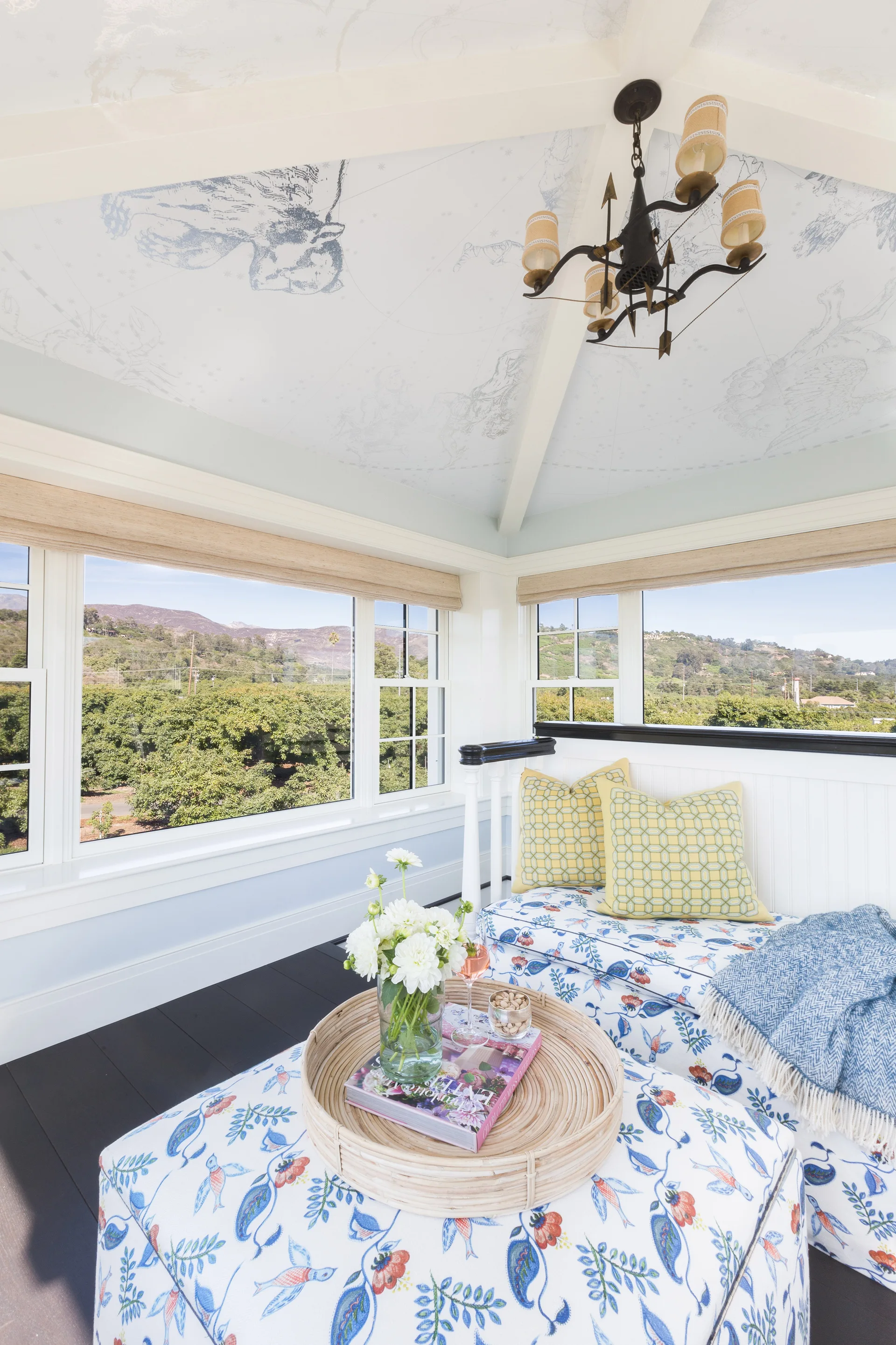 A sunlit sunroom seating area featuring floral-upholstered bench and ottoman, yellow geometric pillows, and a vaulted ceiling with a celestial mural.