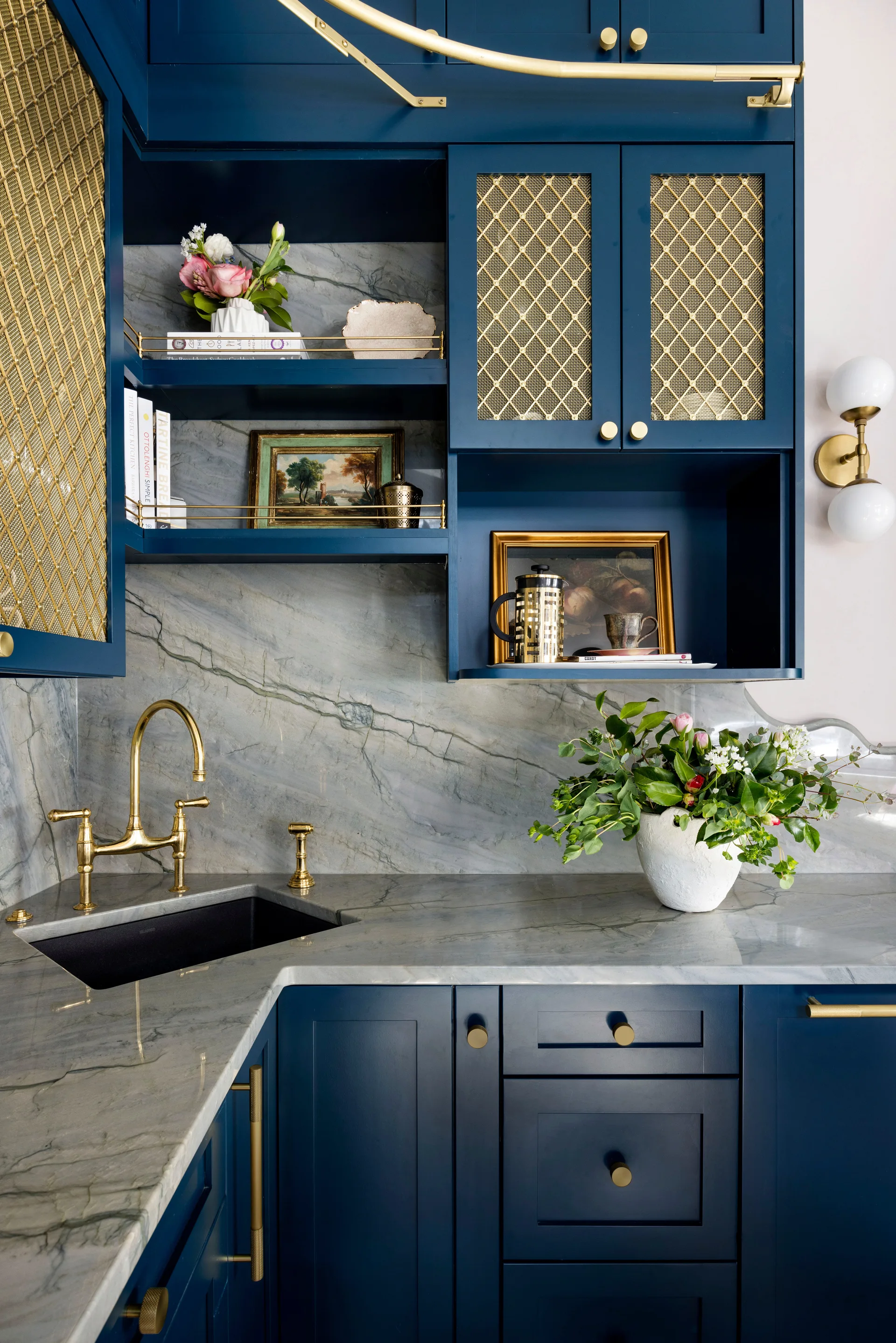 Navy blue kitchen cabinets featuring brass mesh door inserts, a grey veined marble countertop, and a brass bridge faucet over a corner sink.