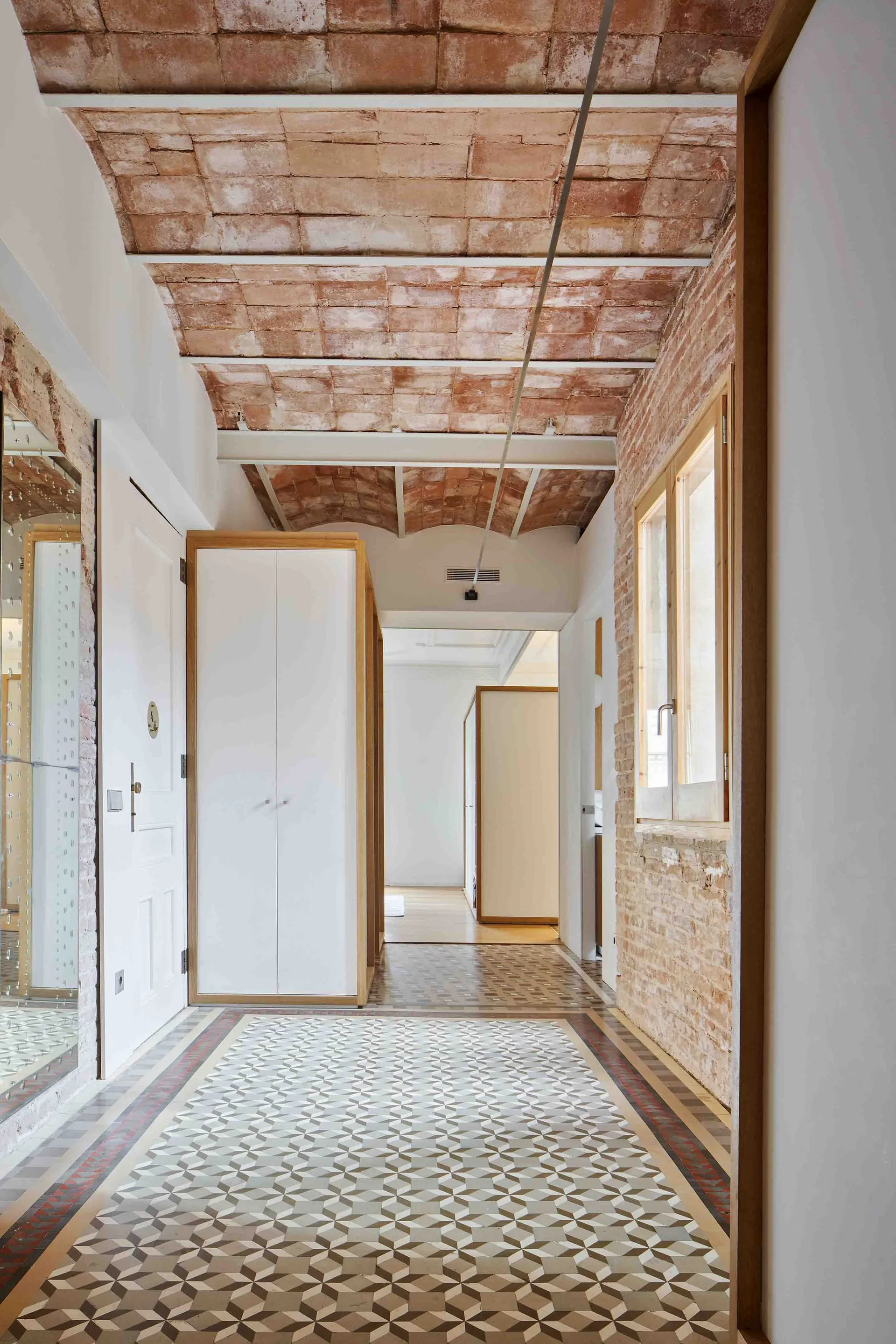 A residential hallway features an exposed terracotta brick vaulted ceiling, geometric patterned floor tiles, and a minimalist white wardrobe with wood trim.