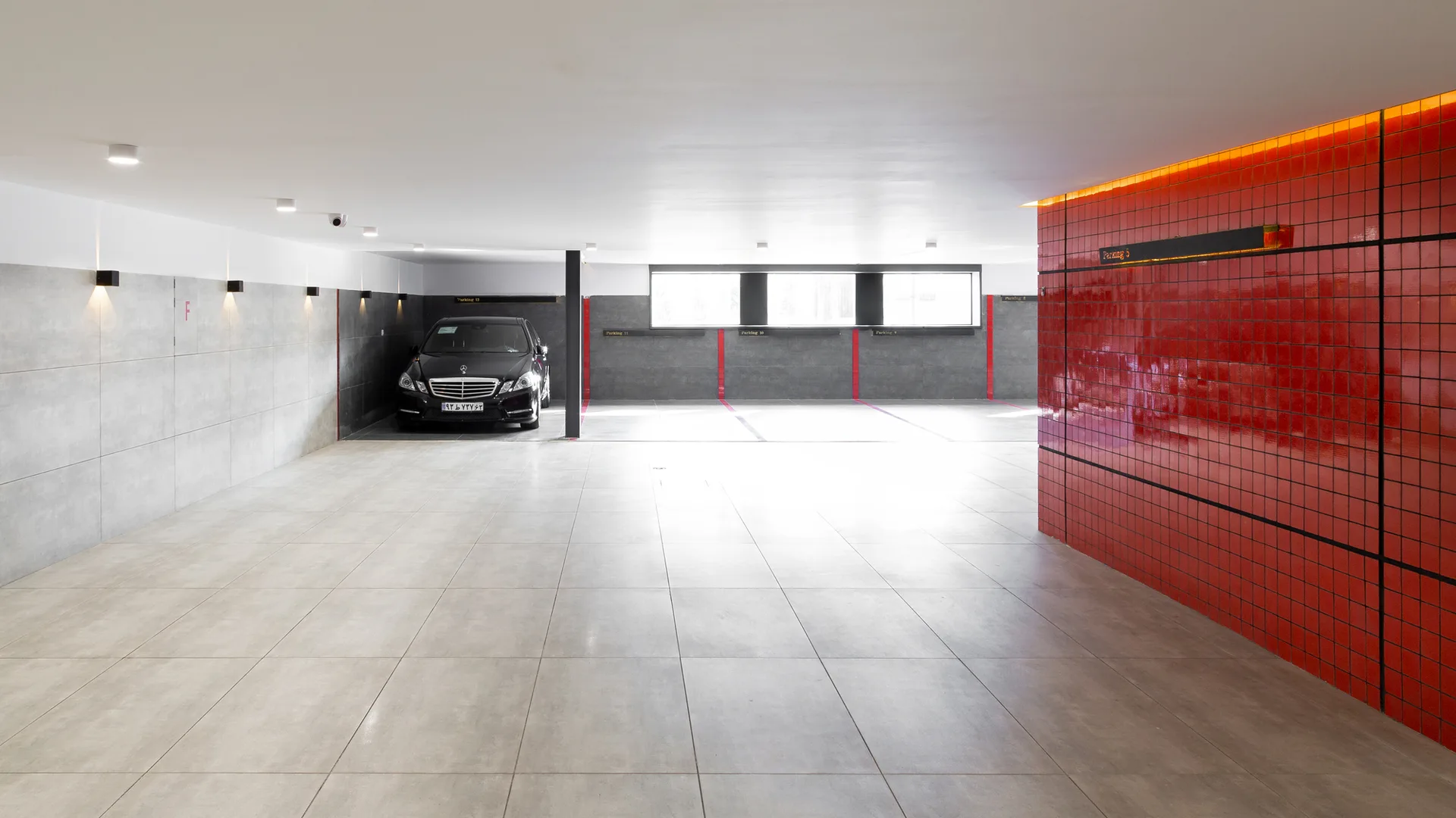 An underground parking garage featuring large grey floor tiles, a parked black car, and a glossy red tiled accent wall.