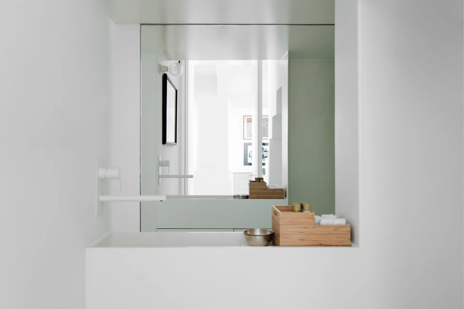 A minimalist white bathroom vanity features an integrated basin, a wall-mounted white faucet, and wooden storage boxes reflecting in a large mirror.