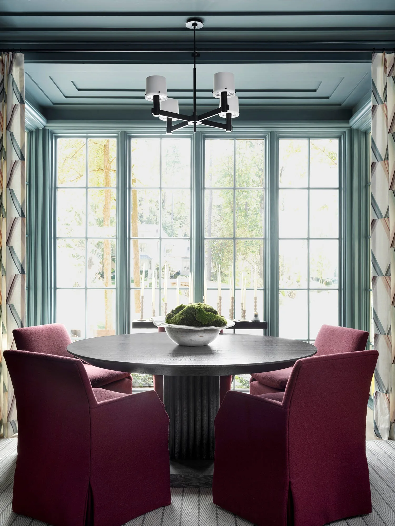 A formal dining room features a round dark wood table and four maroon upholstered chairs set against a large bay window and painted teal walls.