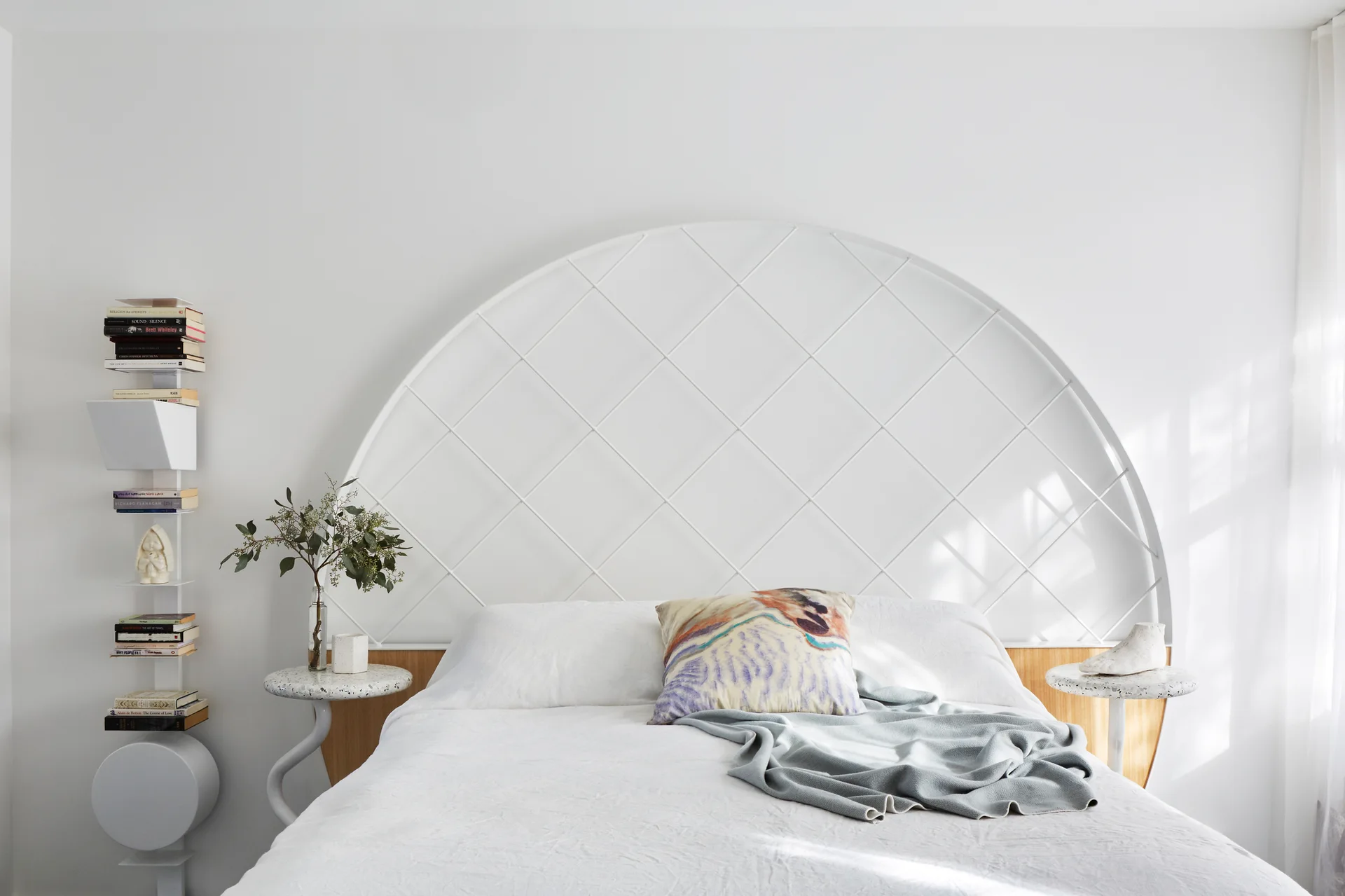 A minimalist white bedroom featuring a bed with a large arched metal grid headboard, matching terrazzo side tables, and a vertical spine bookshelf.