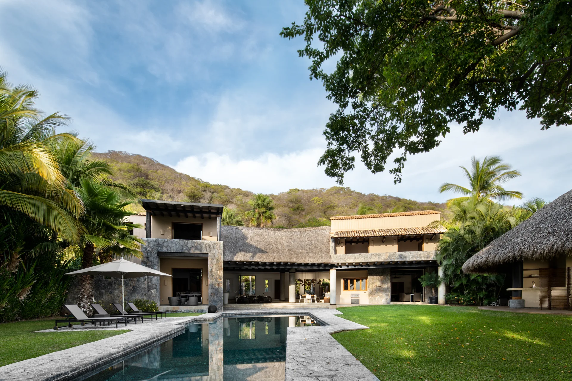 A rectangular swimming pool sits before a two-story tropical villa featuring rough stone walls, a thatched roof, and a manicured green lawn.