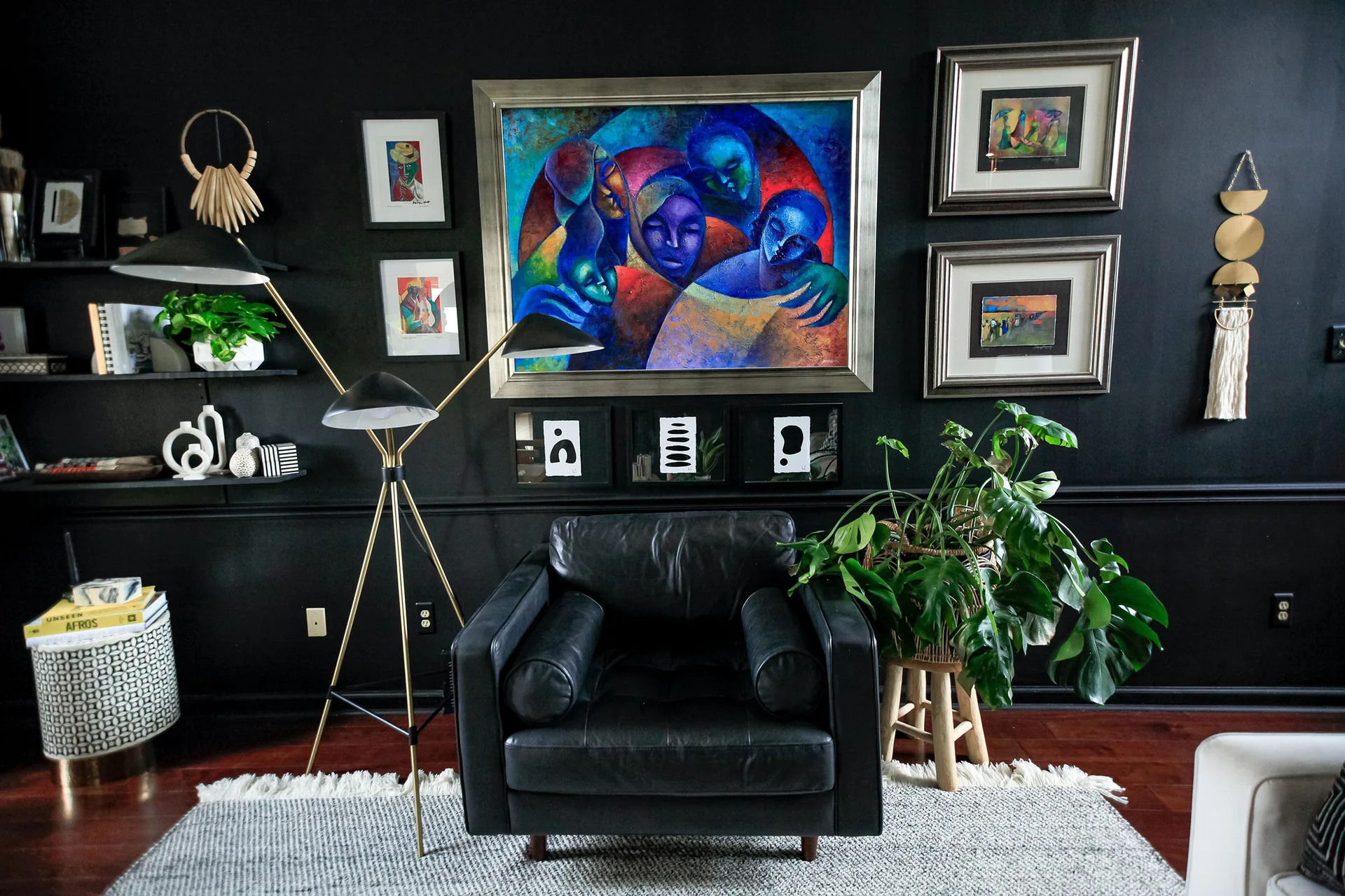A black leather armchair sits in front of a matte black wall decorated with a colorful gallery wall, a gold floor lamp, and a green plant.