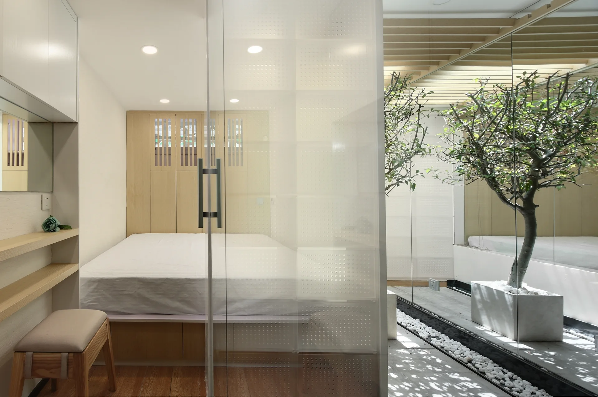 A minimalist bedroom with a platform bed and built-in wood cabinets is separated by a translucent glass partition from an indoor courtyard with a tree.