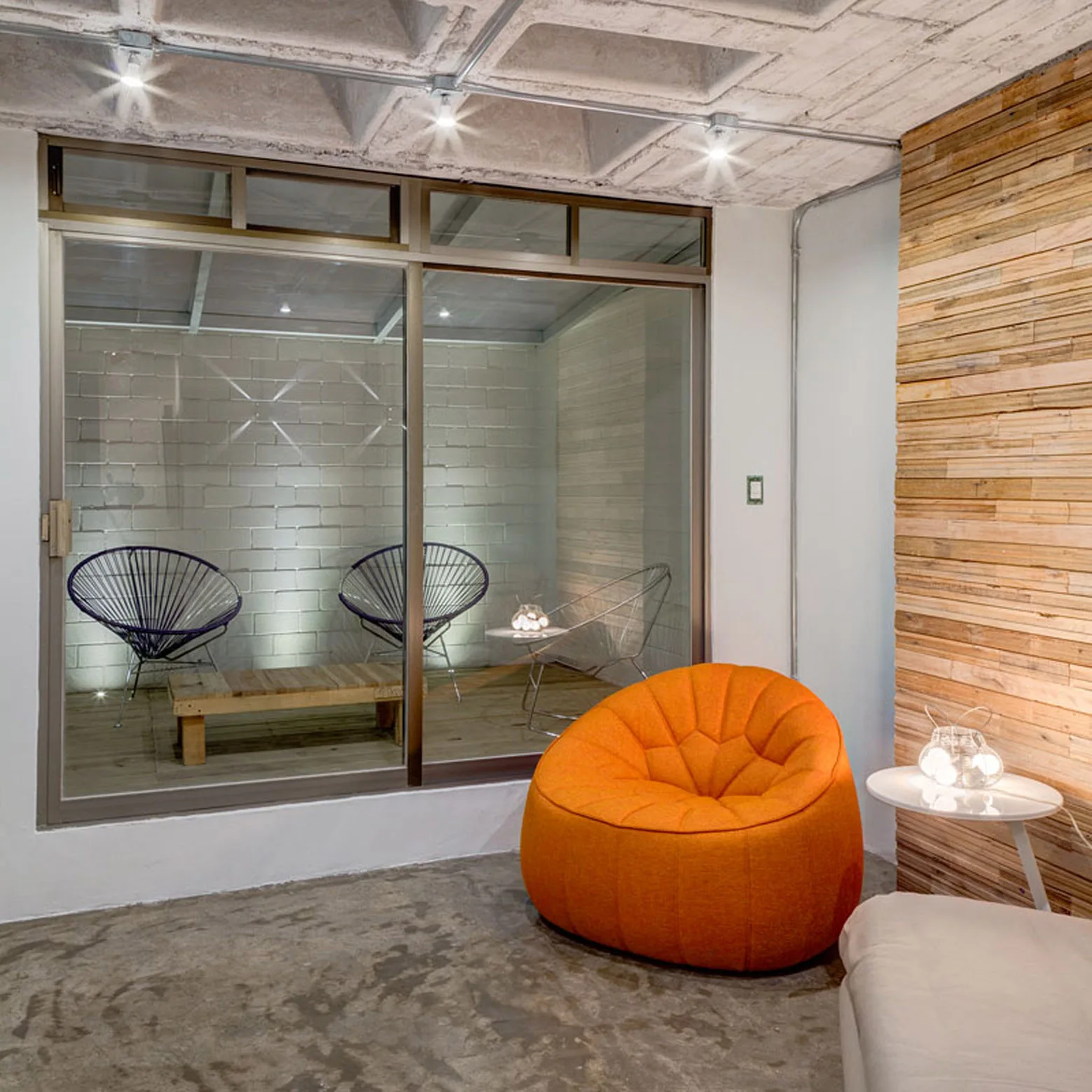 A modern lounge featuring an orange rounded chair, a concrete waffle ceiling, and a glass partition separating two seating areas.