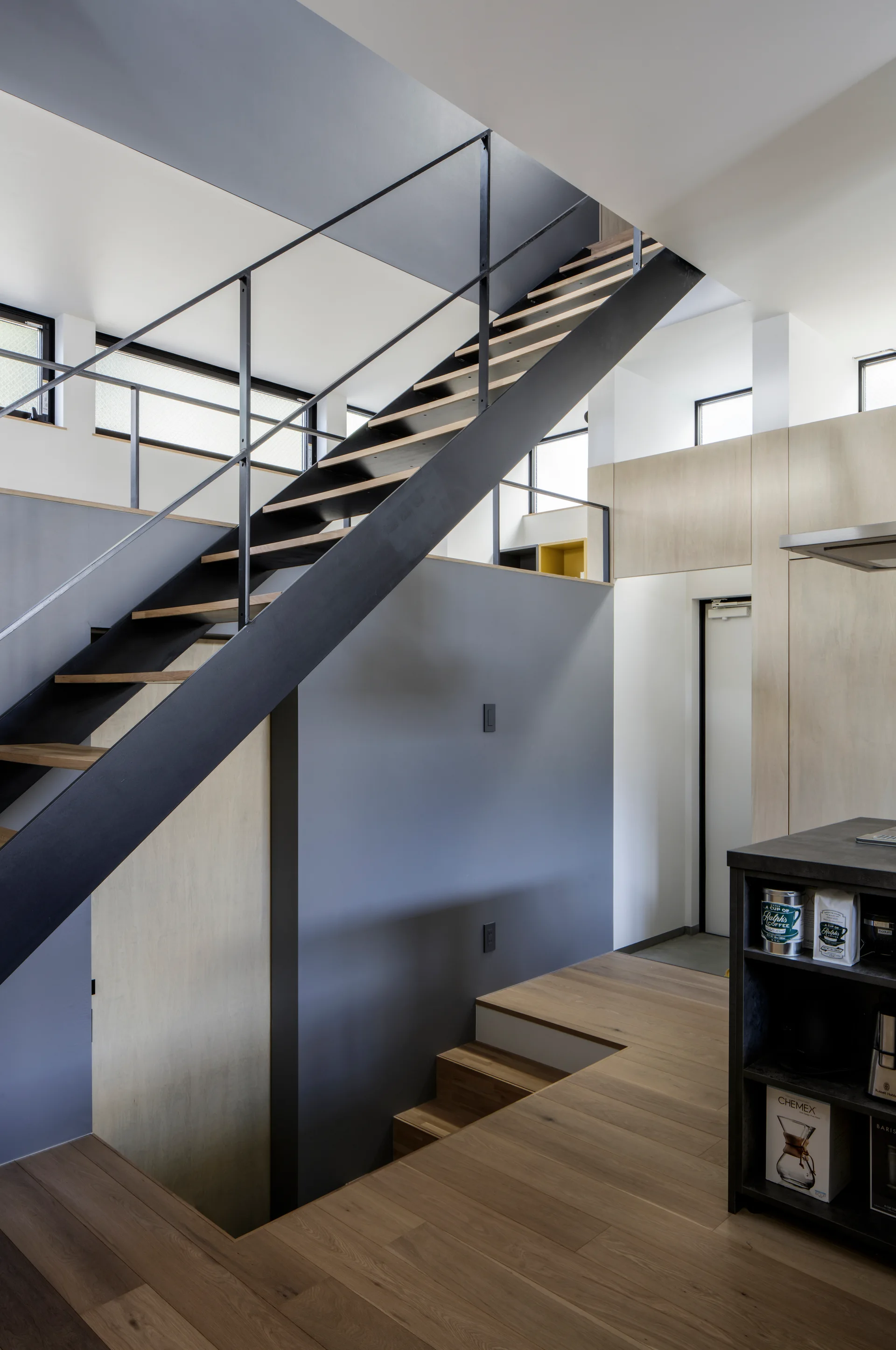 A modern multi-level interior featuring a black steel staircase with wood treads, matte grey walls, and light wood cabinetry near a coffee station.