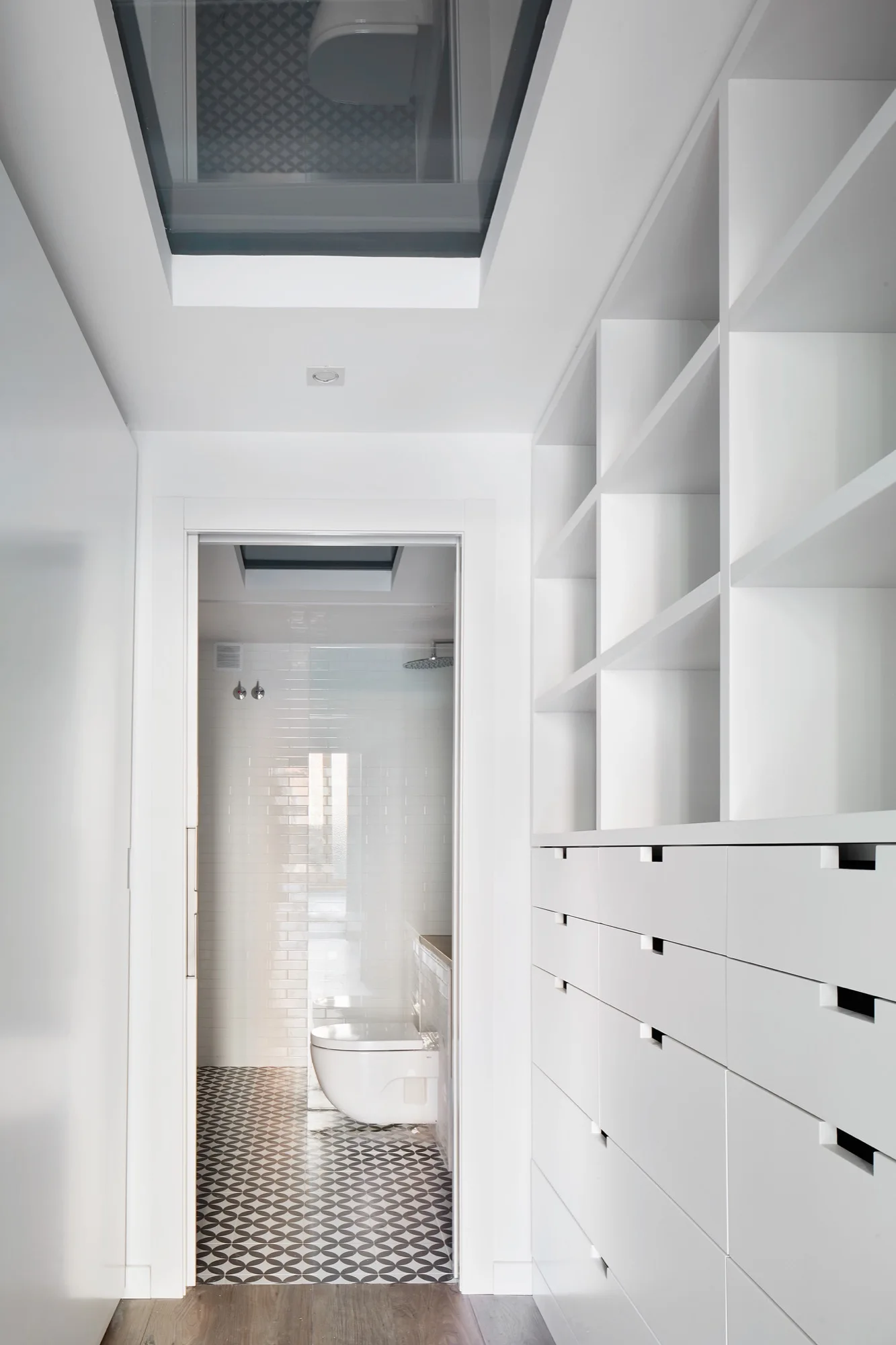 A minimalist white hallway with floor-to-ceiling built-in drawers and shelving leads to a bathroom with geometric tile flooring.