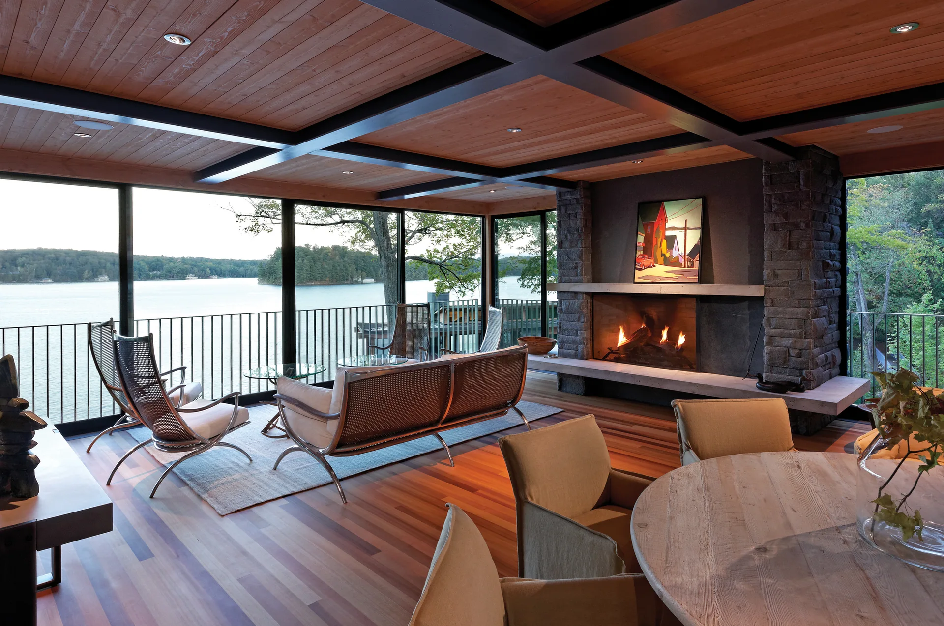 A lakeside living room features glass walls, a stone fireplace, and a wood plank ceiling with dark cross beams.