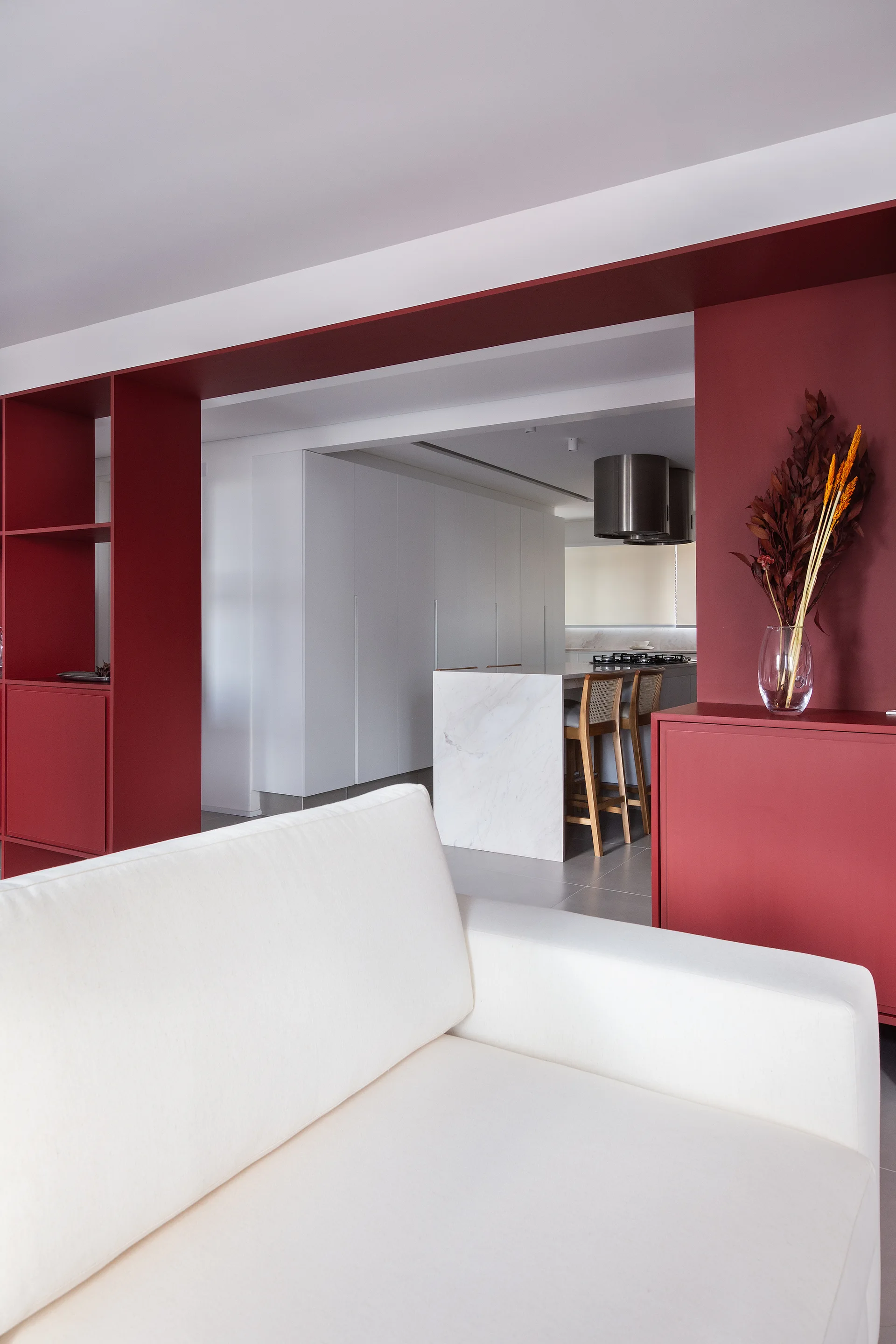 A modern open living space features a white sofa positioned near a bold matte red structural cabinet partition that looks into a minimalist kitchen.