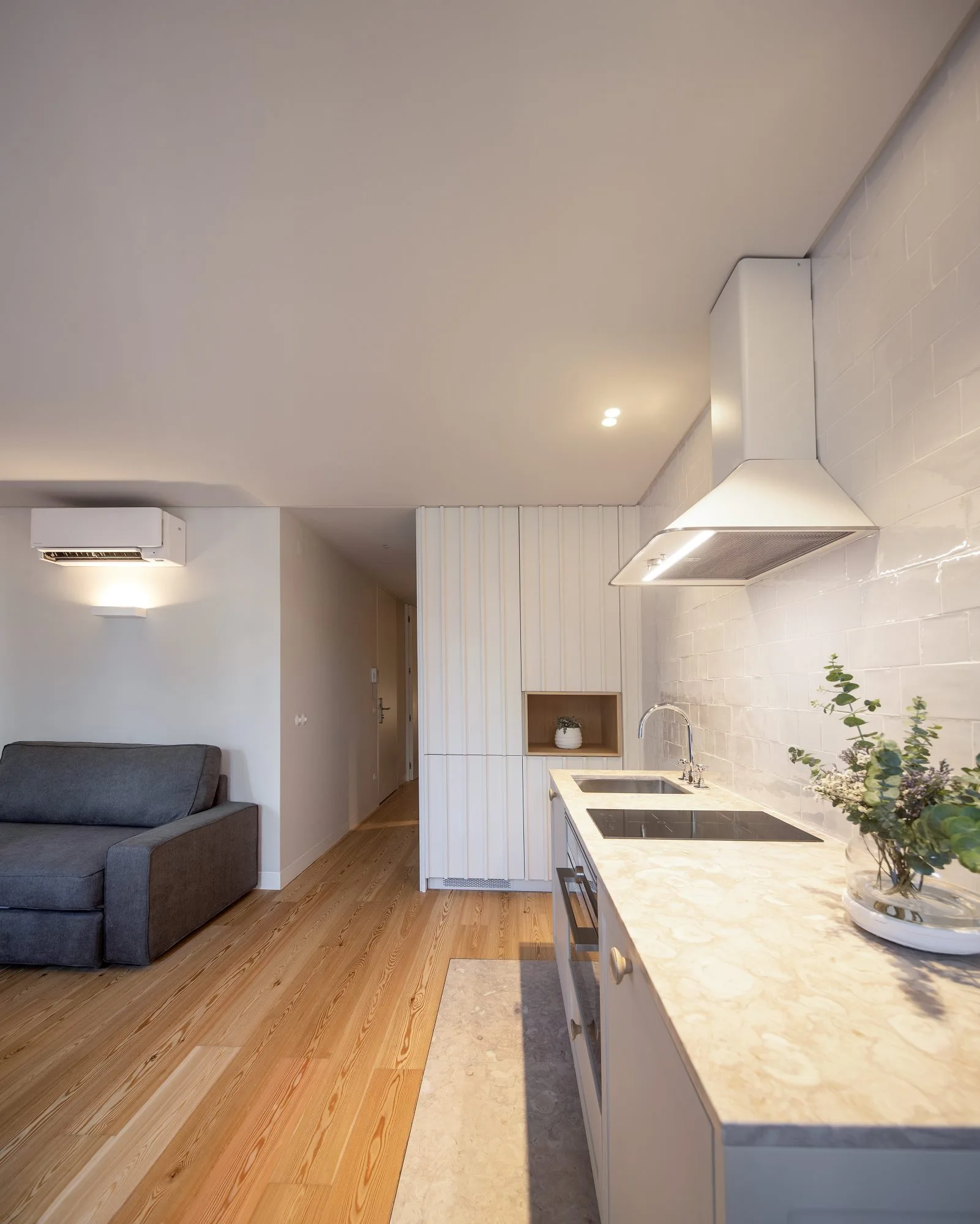 A modern galley kitchen features a stone-like countertop, glossy subway tile backsplash, and white vertical slatted cabinets next to a grey sofa.