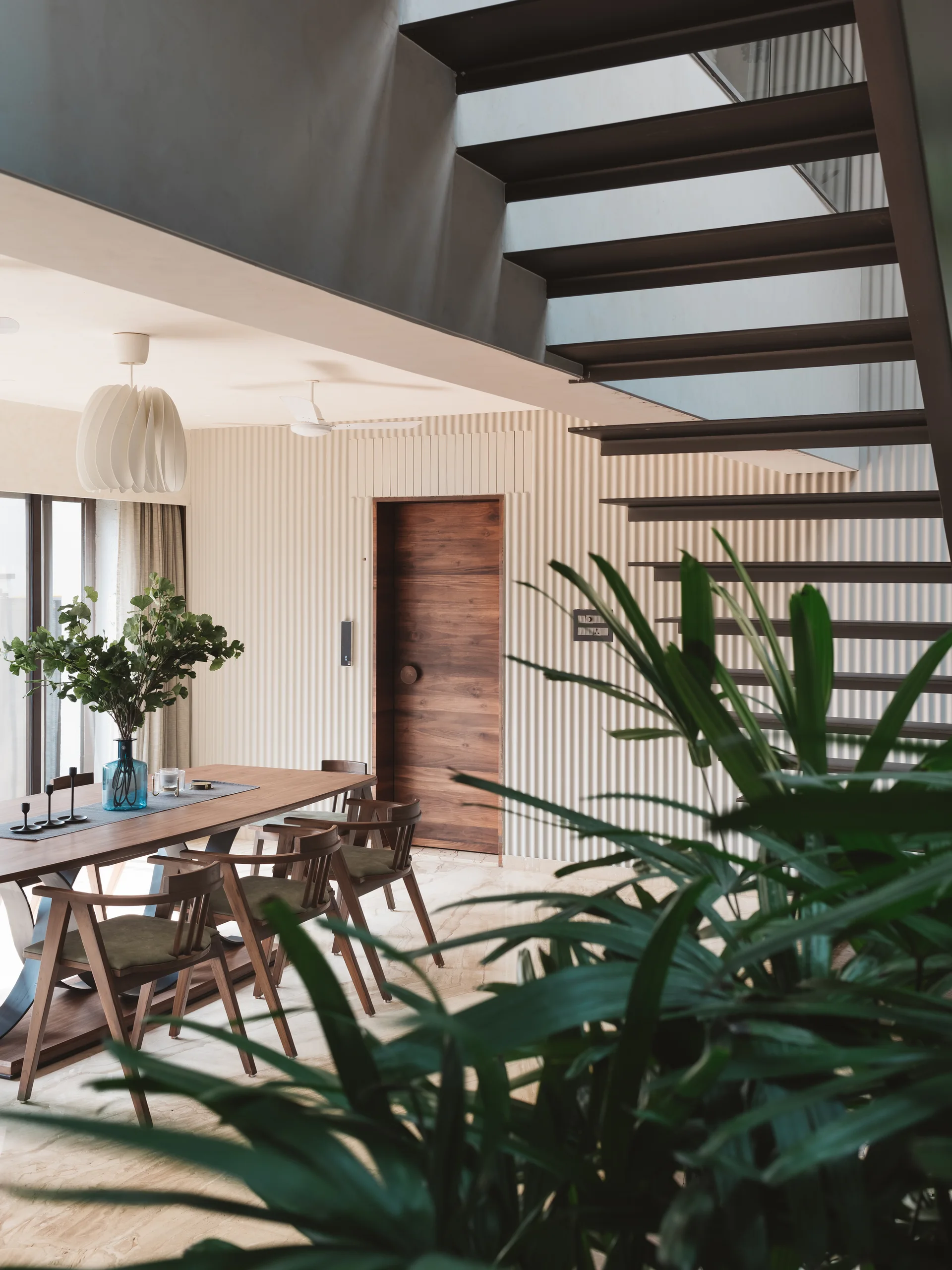 A modern dining area featuring a wooden table and chairs, a cream vertically ribbed wall with a flush door, and a dark metal floating staircase.