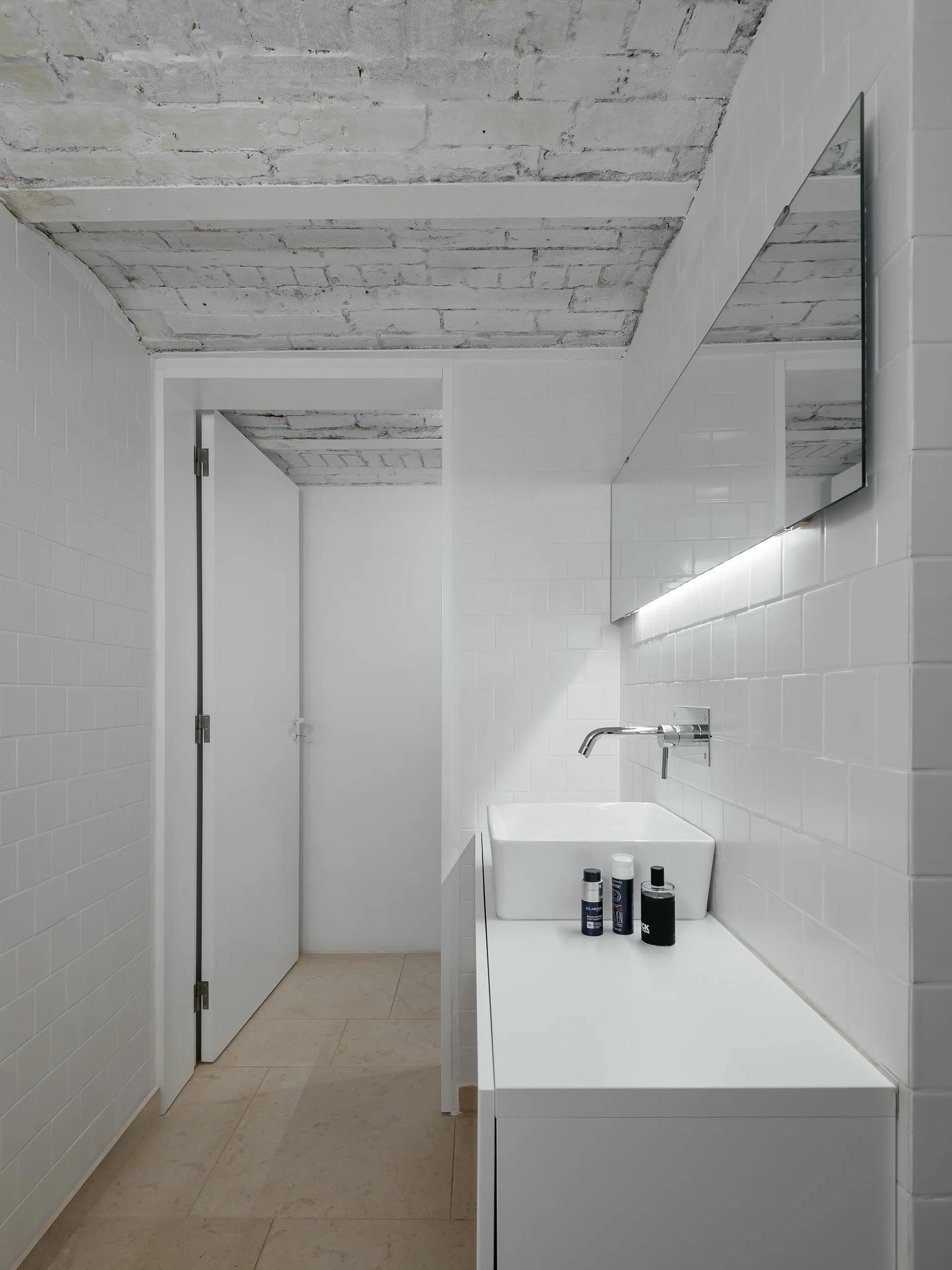 A minimalist white bathroom features square tiled walls, a rectangular vessel sink on a white vanity, and a rustic painted brick ceiling.
