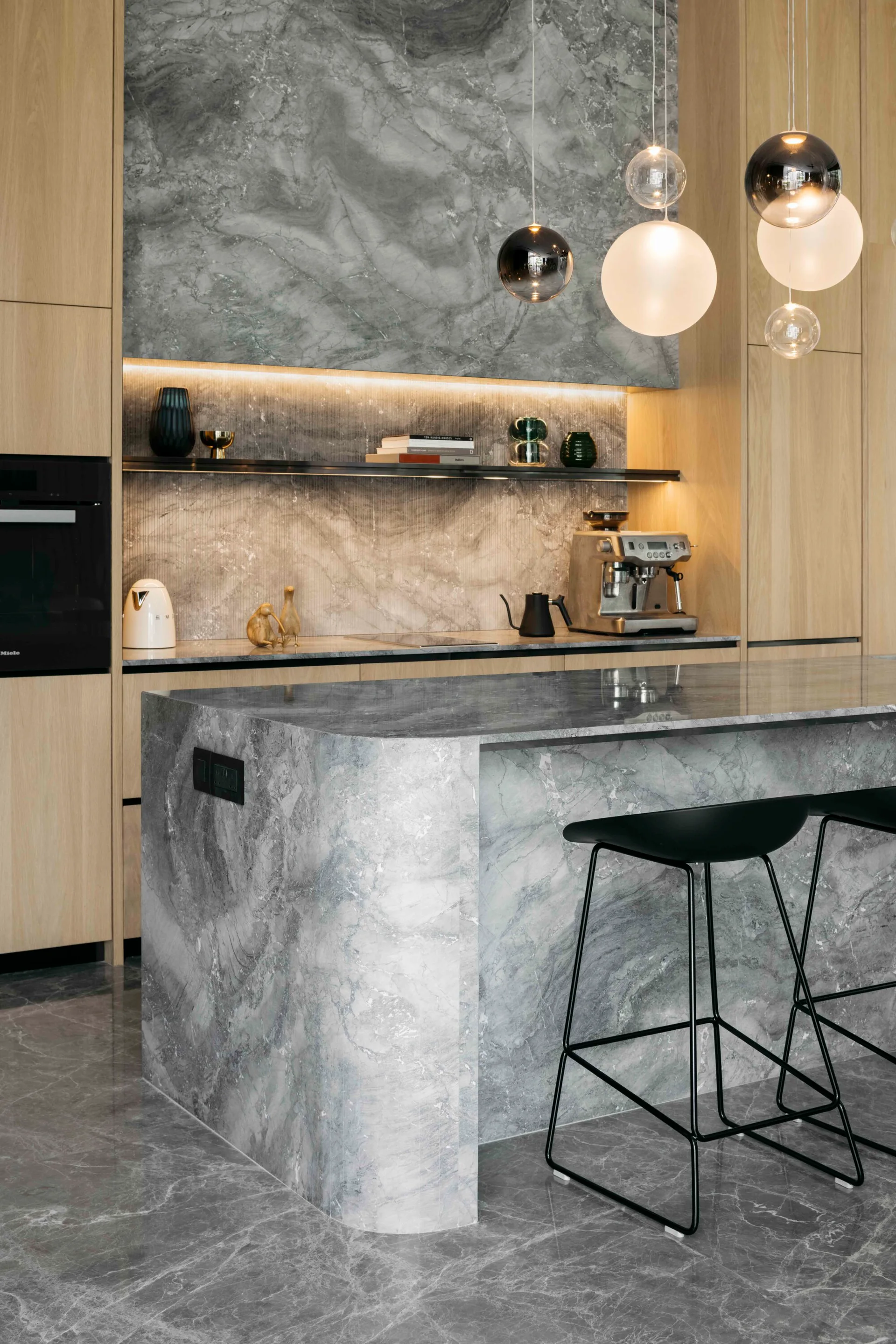 A modern kitchen featuring a curved grey stone island with black barstools, wood cabinetry, and suspended orb pendant lights.