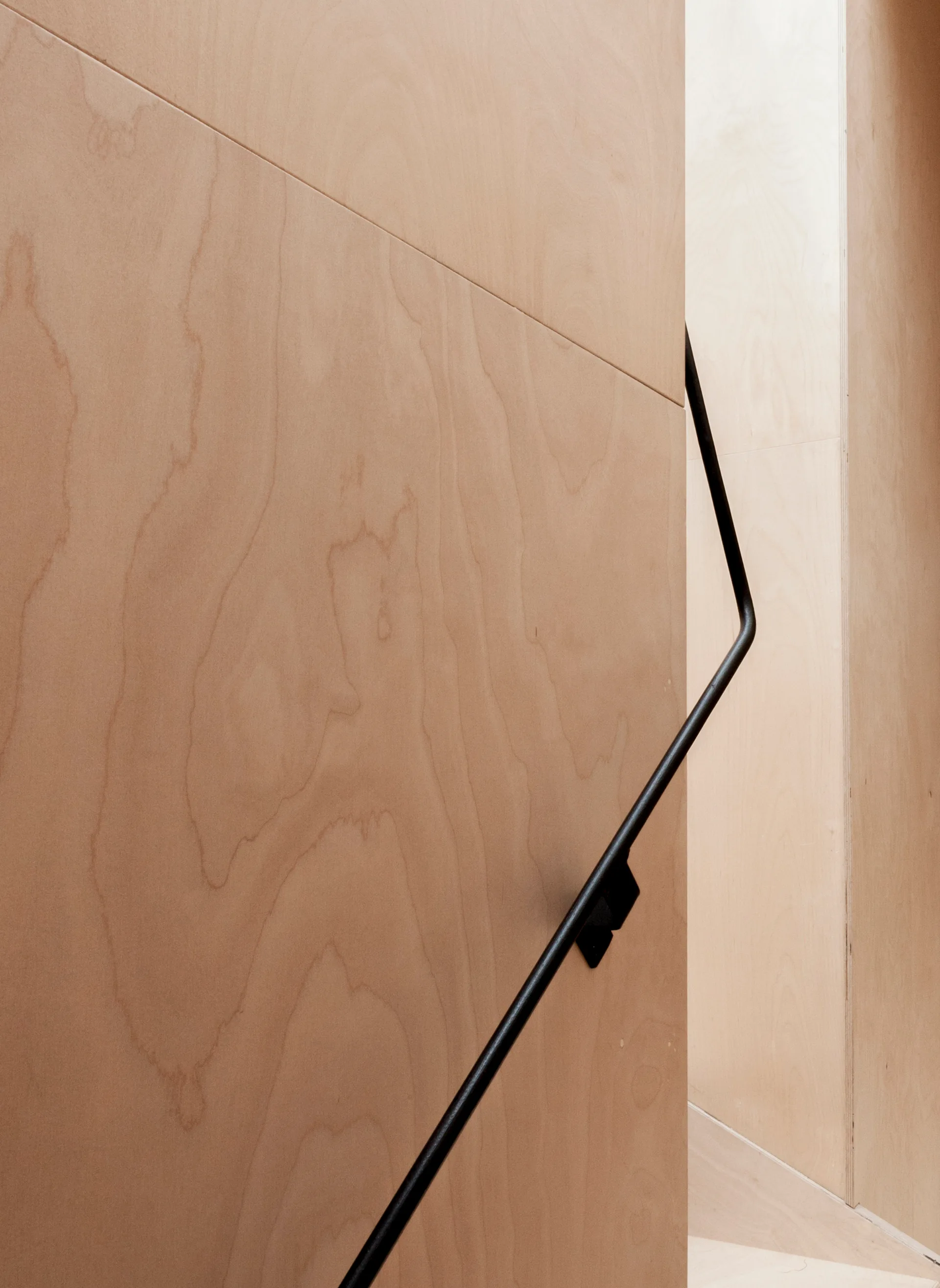 A minimalist staircase featuring light-toned plywood wall paneling and a slim, black metal handrail.