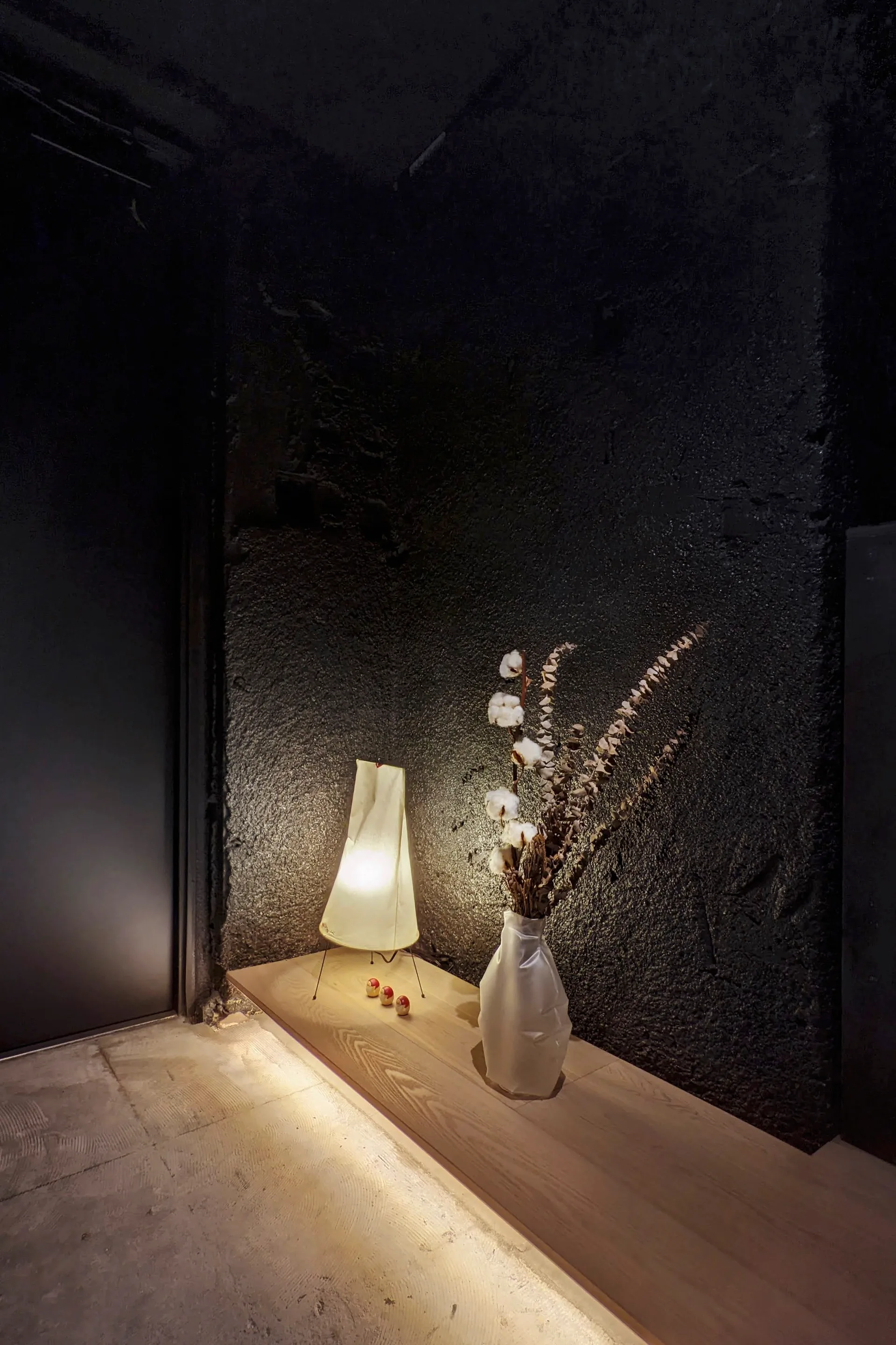 A low wood floating shelf decorated with a glowing paper lamp and dried cotton branches in a white vase against a dark textured wall.
