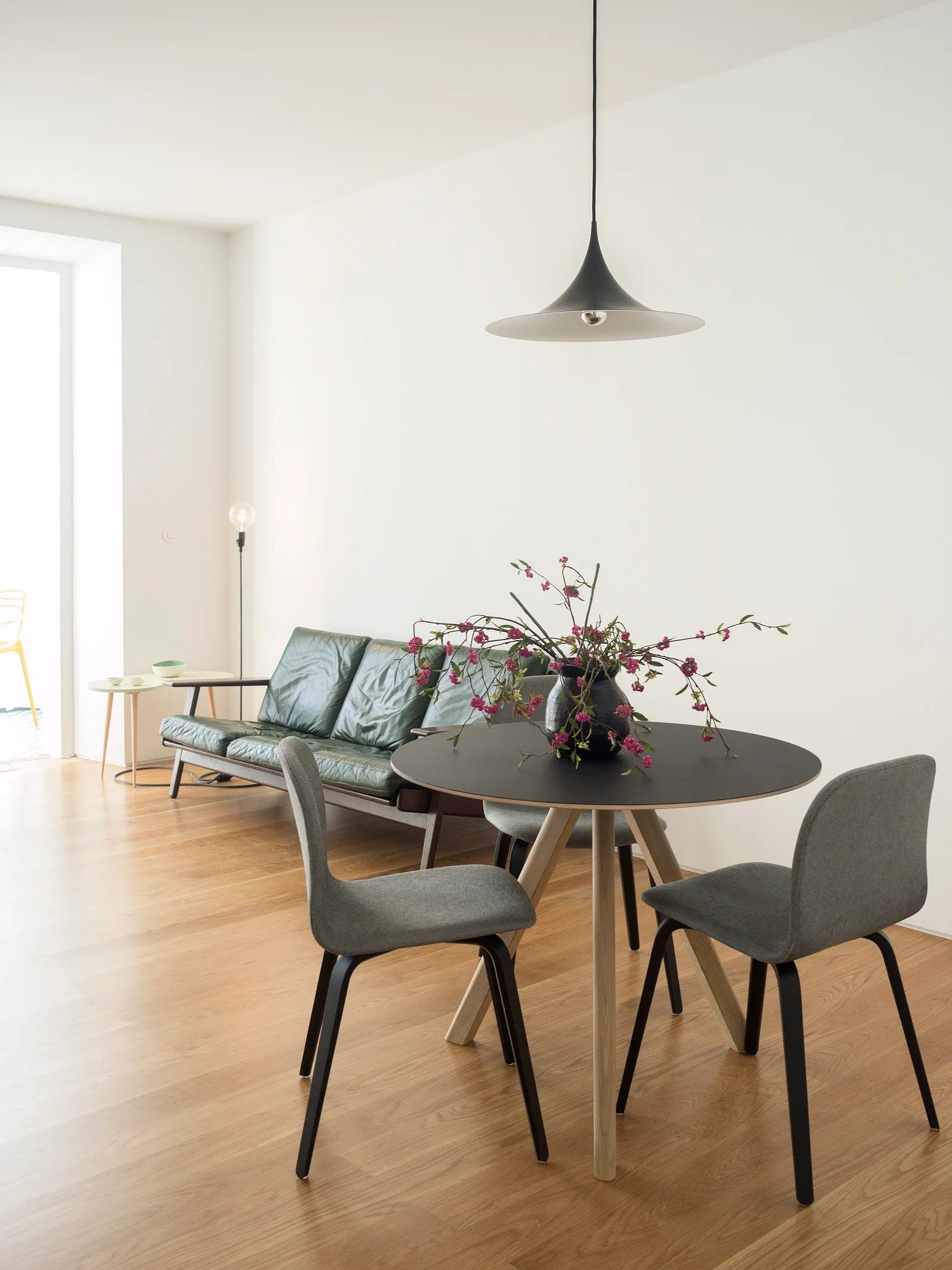 A minimalist dining area with a round black table, grey chairs, and a dark green leather sofa against white walls.