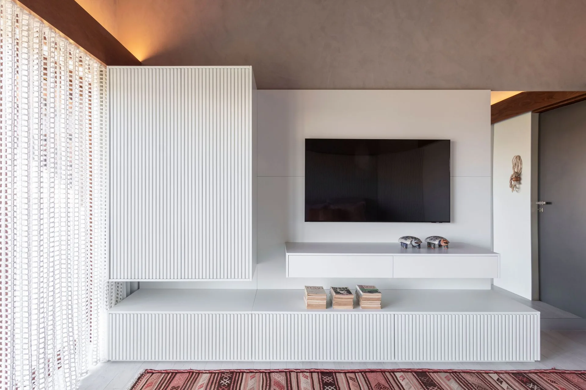 A minimalist living room features a white slatted built-in media console with a wall-mounted television, a grey ceiling, and a patterned rug.