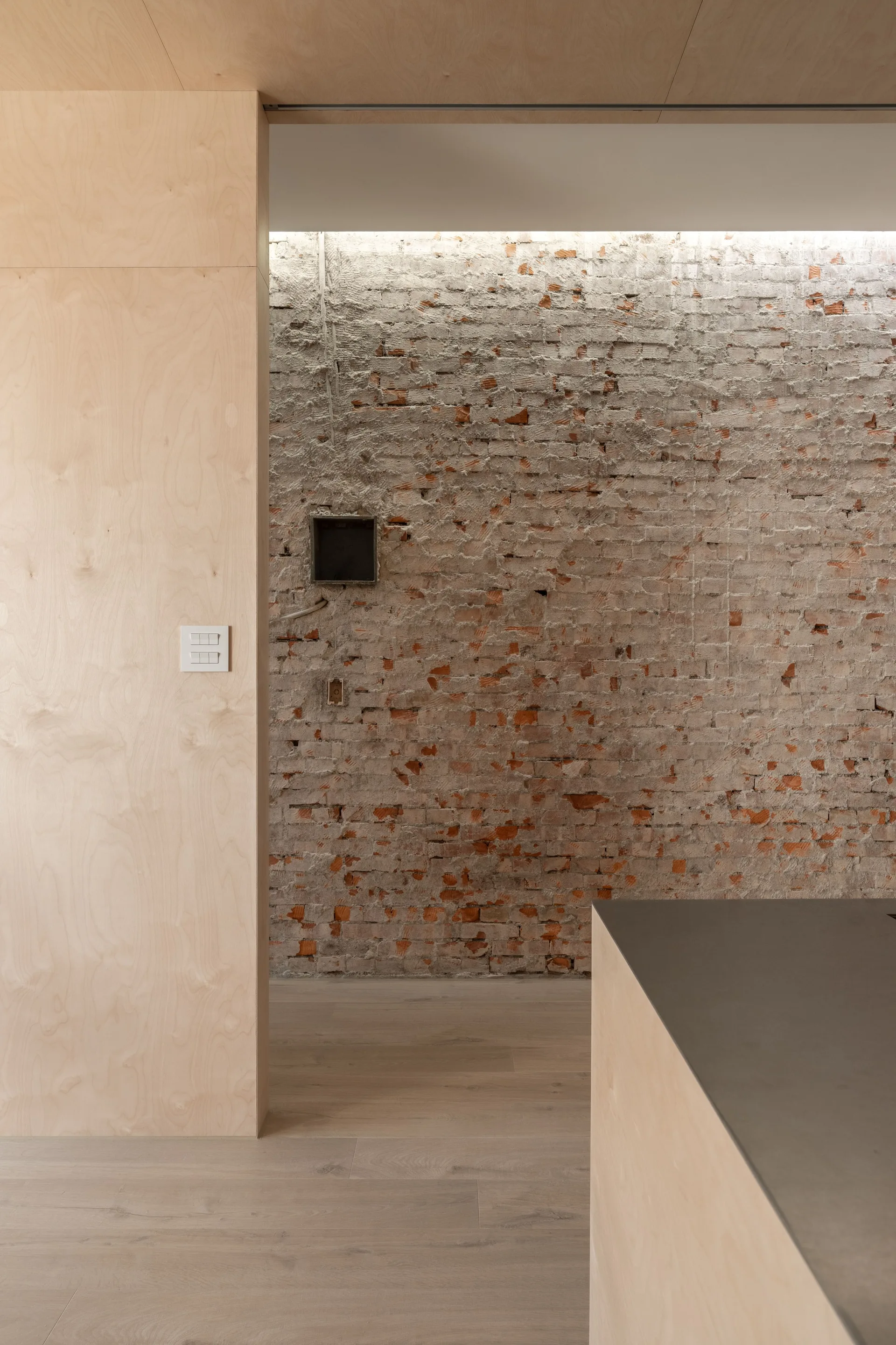 An interior view showing a textured, weathered brick wall adjacent to smooth light wood plywood paneling under a recessed warm cove light strip.