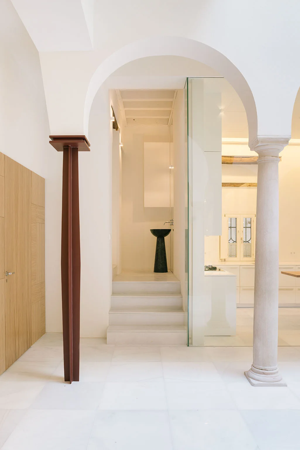 A bright white hallway features marble flooring, a stone column, a brown metal I-beam supporting an arch, and steps leading to a dark pedestal sink.