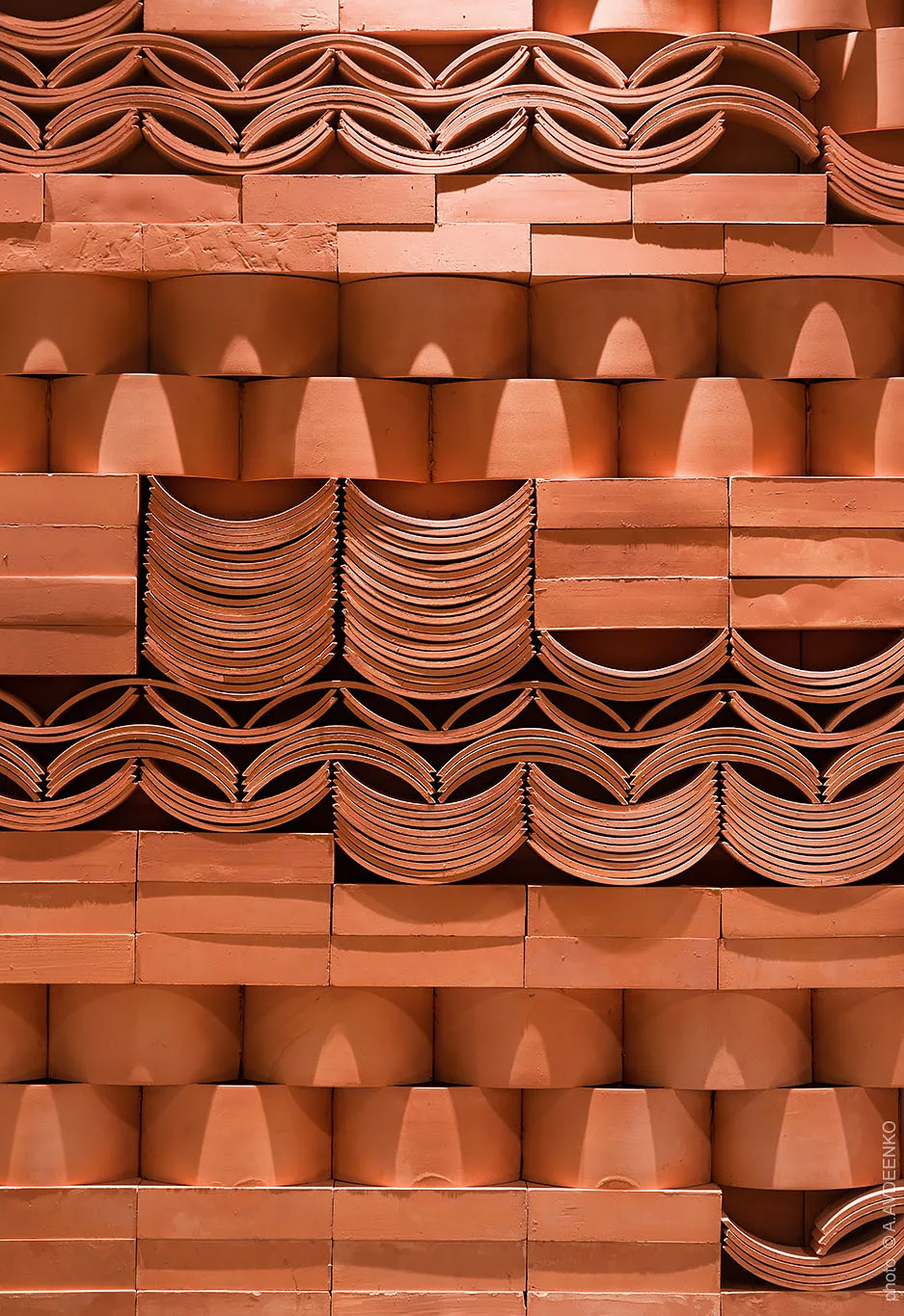 A close-up view of a wall featuring stacked terracotta clay tiles arranged in a repetitive geometric pattern with curved and rectangular shapes.