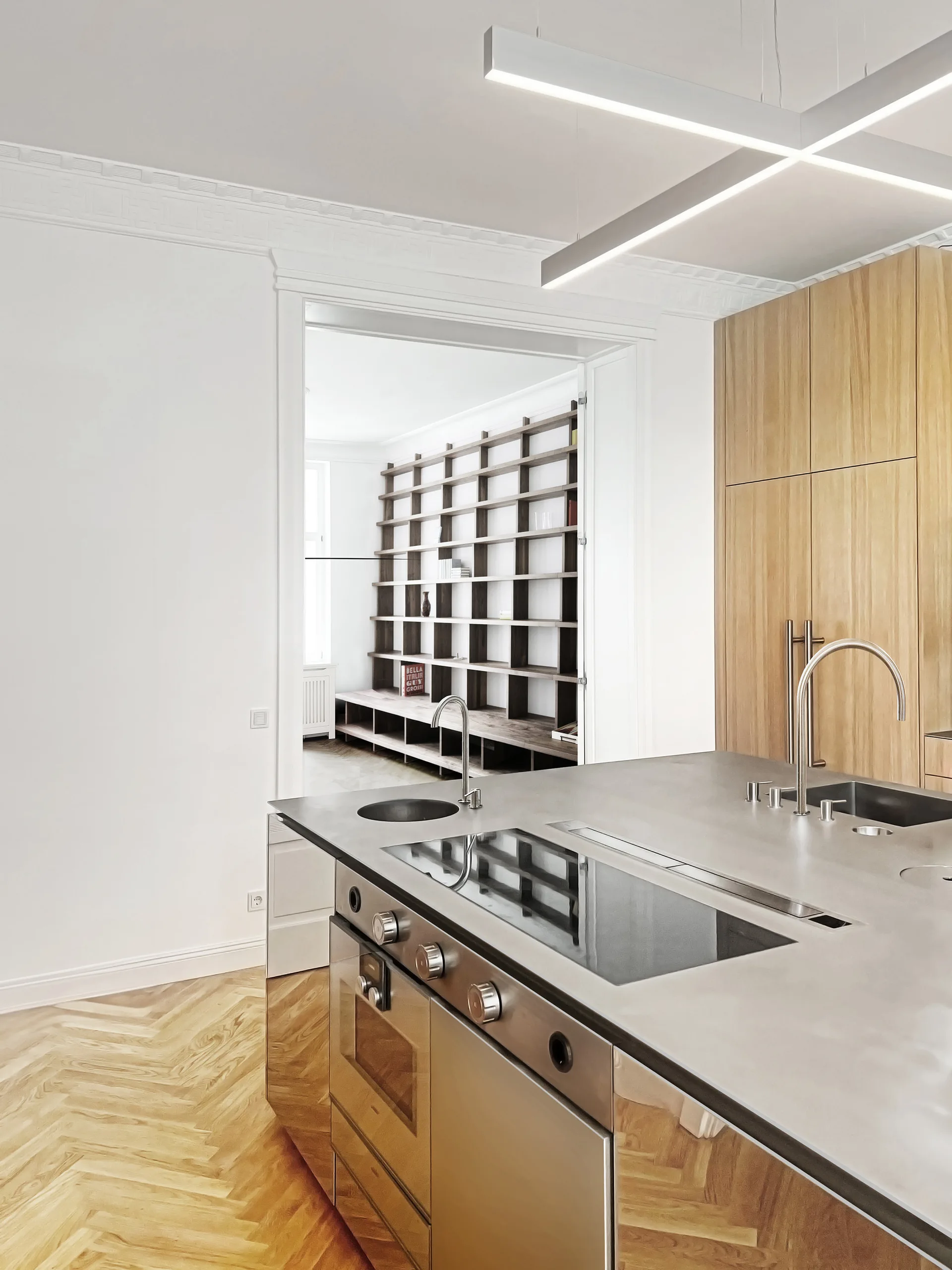 A modern kitchen with a stainless steel island and light wood cabinets, looking through a doorway toward a dark wood shelving unit in the next room.