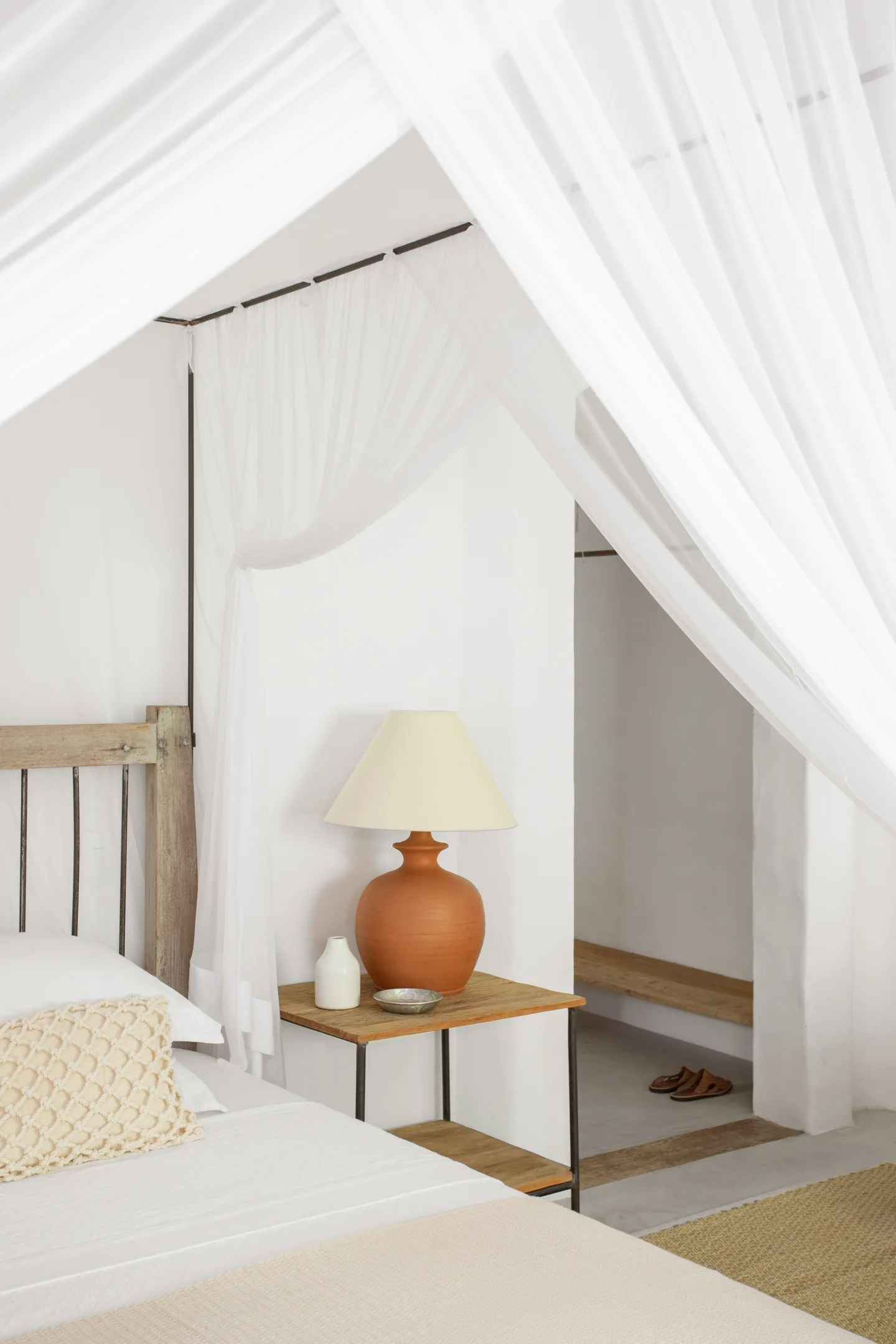 A rustic wooden four-poster bed with white fabric drapes stands next to a wooden nightstand and a terracotta table lamp.