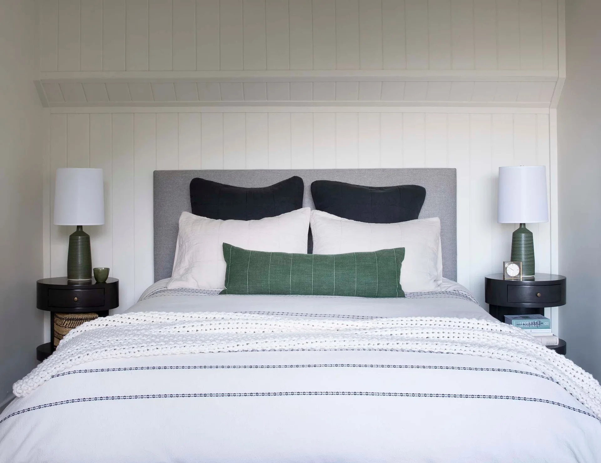 Symmetrical bedroom featuring a bed with a grey headboard, a green lumbar pillow, and matching dark round nightstands with green lamps.