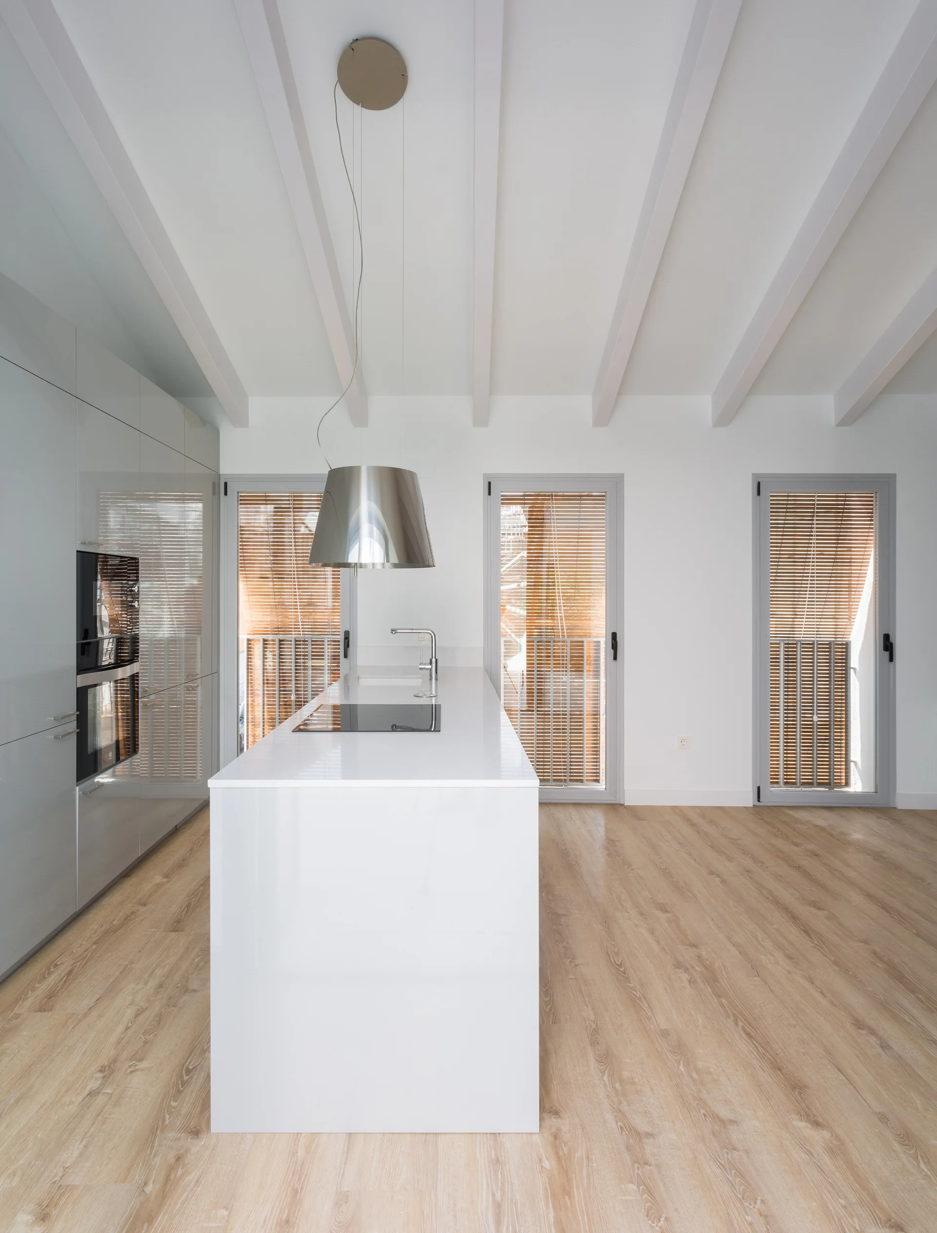 A minimalist white kitchen featuring a central island, glossy cabinetry, white ceiling beams, and three doors with light wood venetian blinds.